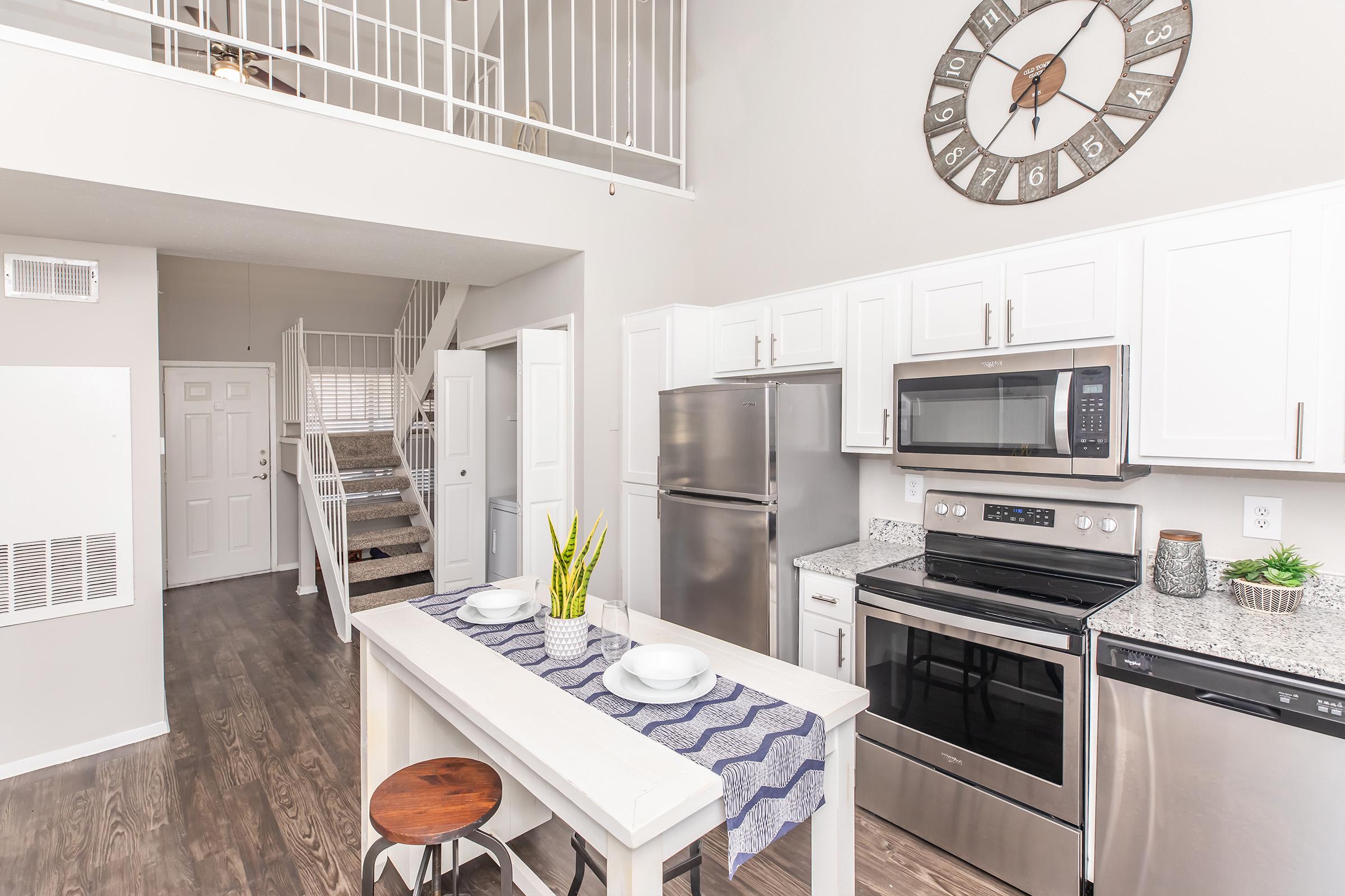 Modern kitchen and dining area featuring stainless steel appliances, a granite countertop, white cabinetry, and a decorative wall clock. The space includes a table set for two and opens to a staircase leading to an upper level. The overall design has a bright and airy feel with light-colored walls and ample natural light.