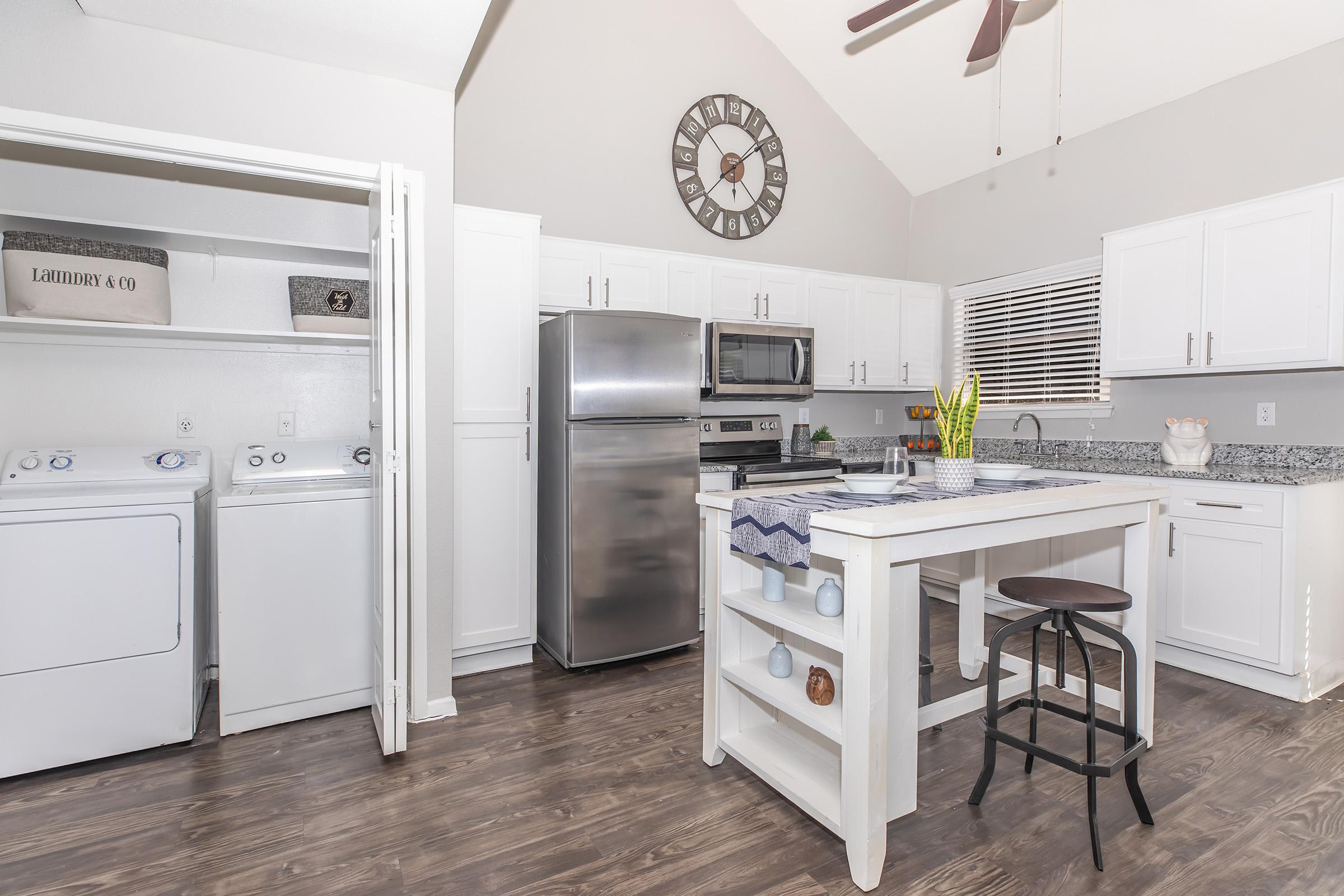 A modern kitchen with white cabinetry and gray walls. Stainless steel appliances, including a fridge and microwave, are visible. A central island with a dark stool and decorative items on top adds style. To the side, there's a laundry area with washer and dryer units. Natural light brightens the space.