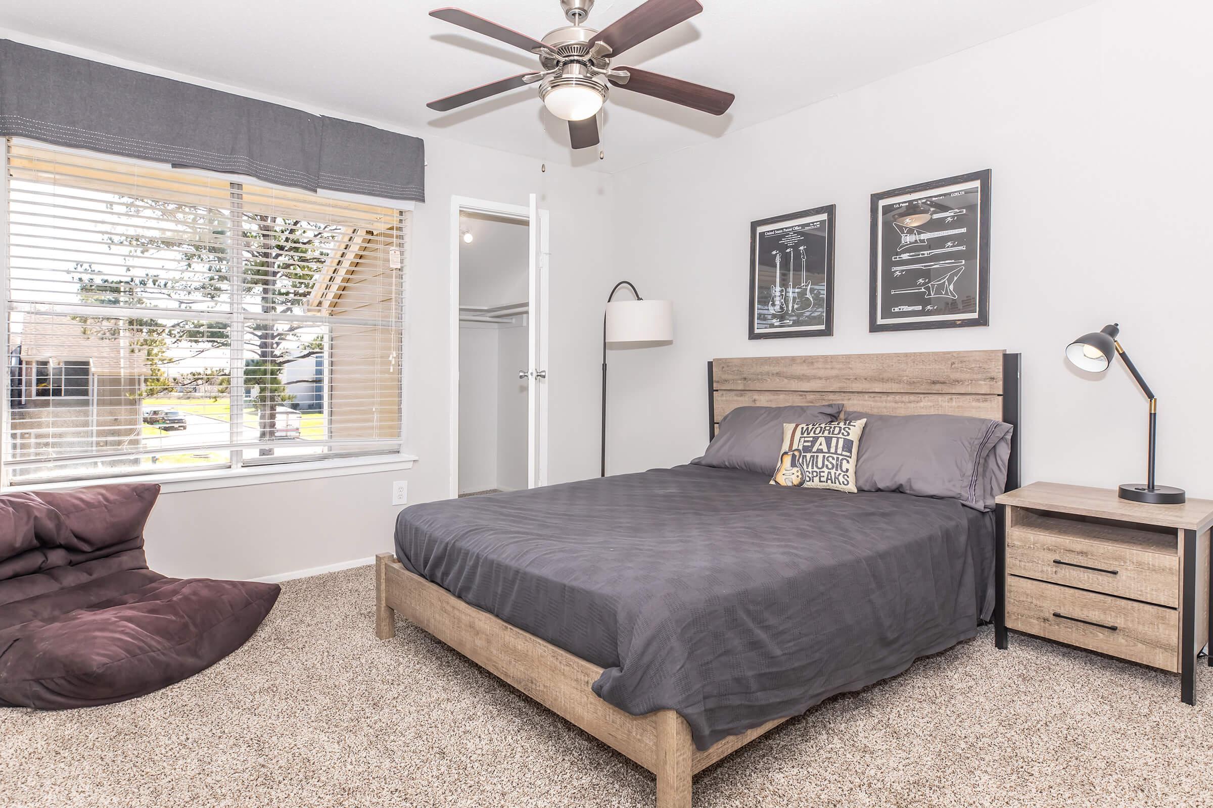 A cozy bedroom featuring a wooden bed frame with gray bedding, a bedside table with a lamp, and a plush bean bag chair on carpeted flooring. Natural light streams through a window with blinds, and two framed prints hang on the wall above the bed. A ceiling fan adds to the comfortable atmosphere.