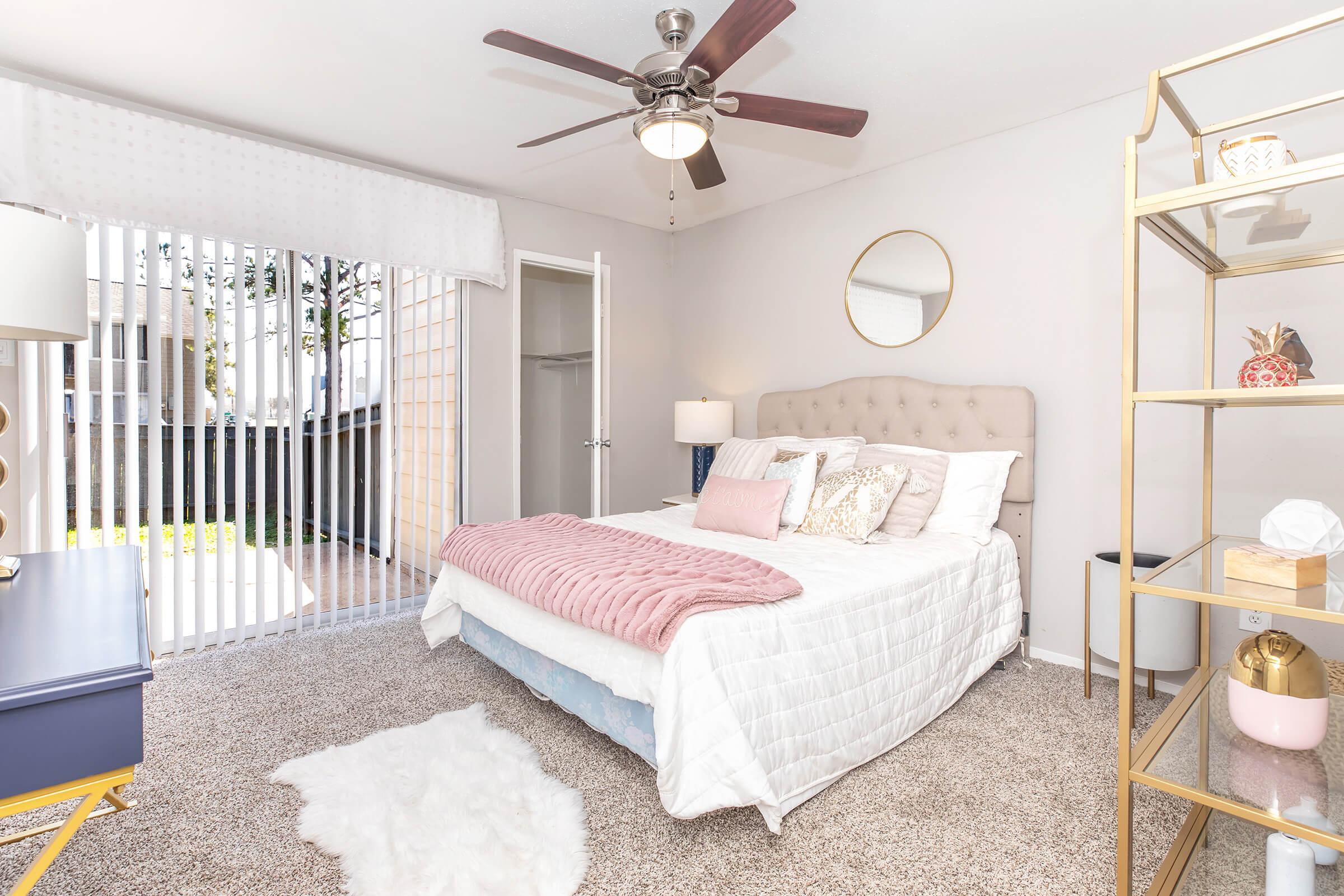 A bright, modern bedroom featuring a queen-sized bed with a plush pink throw, decorative pillows, and a cozy rug. There is a ceiling fan overhead, a stylish gold shelf display with decorative items, and large sliding doors leading to an outdoor space with greenery. Natural light fills the room, creating a warm atmosphere.