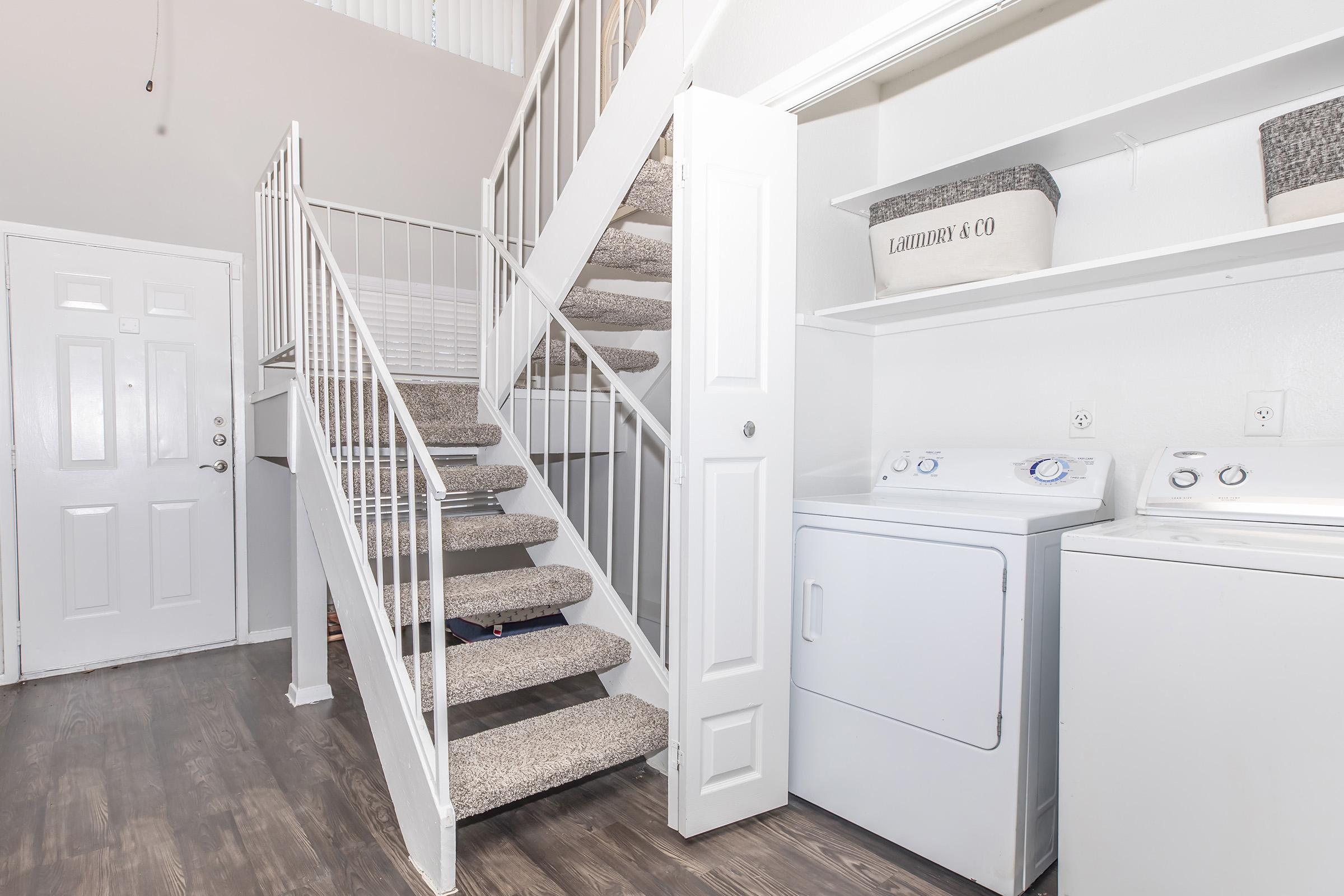 A well-lit interior space featuring a staircase with white railings leading to an upper level. On one side is a laundry area with a washer and dryer, and on the other side is a door leading outside. The walls are painted in light colors, and the flooring is a wood-like laminate, creating a clean and modern look.