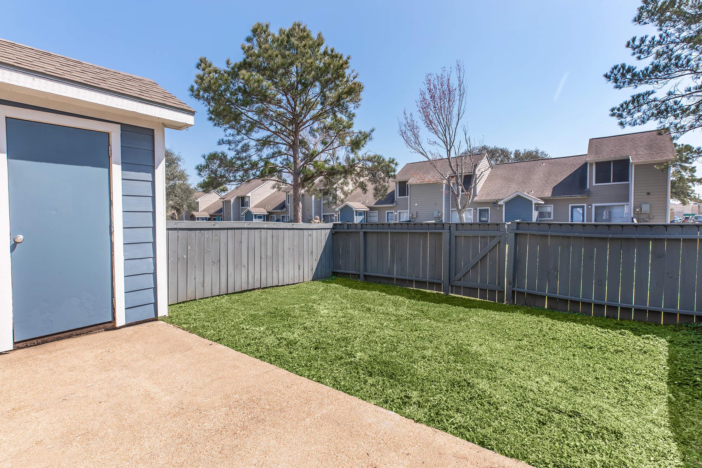 A small fenced backyard featuring green artificial grass, a concrete patio area, and a blue shed with a door. In the background, there are residential buildings with balconies and trees nearby under a clear blue sky.