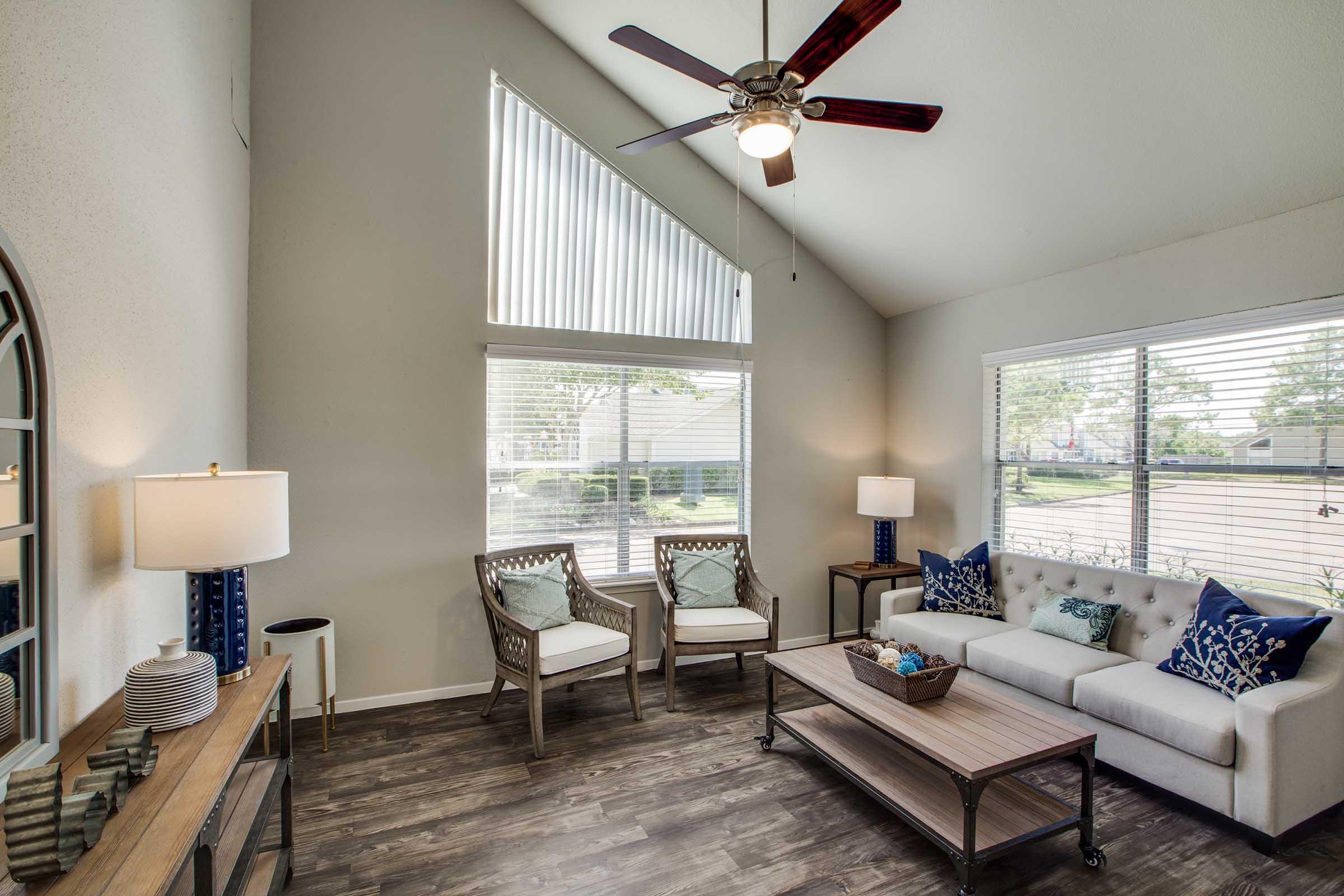 A bright and airy living room featuring a light-colored sofa with decorative blue pillows, two armchairs with matching pillows, a wooden coffee table, and a stylish lamp. Large windows let in natural light, and the room has a modern ceiling fan and tasteful decor elements, creating a welcoming atmosphere.