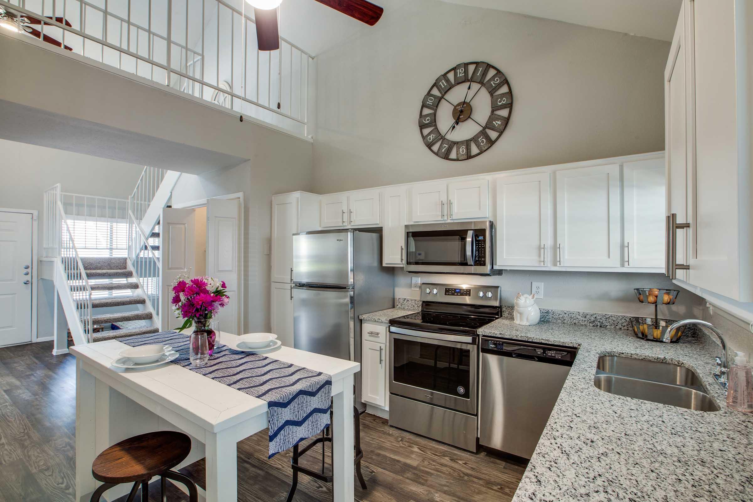 A modern kitchen featuring white cabinetry, stainless steel appliances, and a granite countertop. A round table set for two with a vase of pink flowers stands in the center. A wall clock is prominently displayed above the kitchen area, and a staircase leading to an upper level is visible in the background.