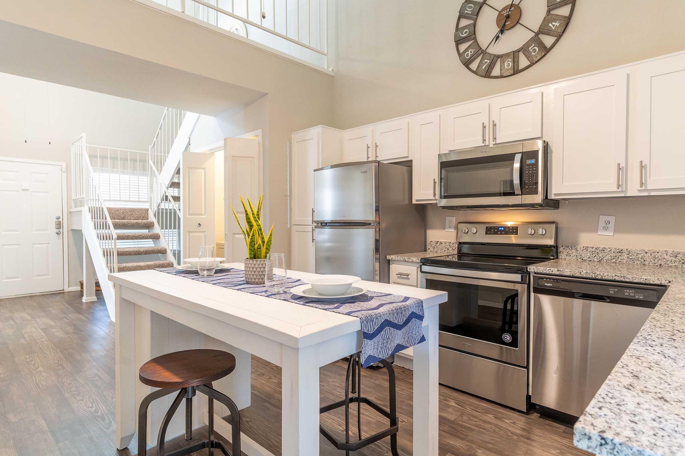 Modern kitchen with white cabinets, stainless steel appliances, and granite countertops. A small dining table with a blue table runner and two stools is centered, complemented by a decorative plant. The space is well-lit, featuring a clock on the wall and stairs leading to an upper level.