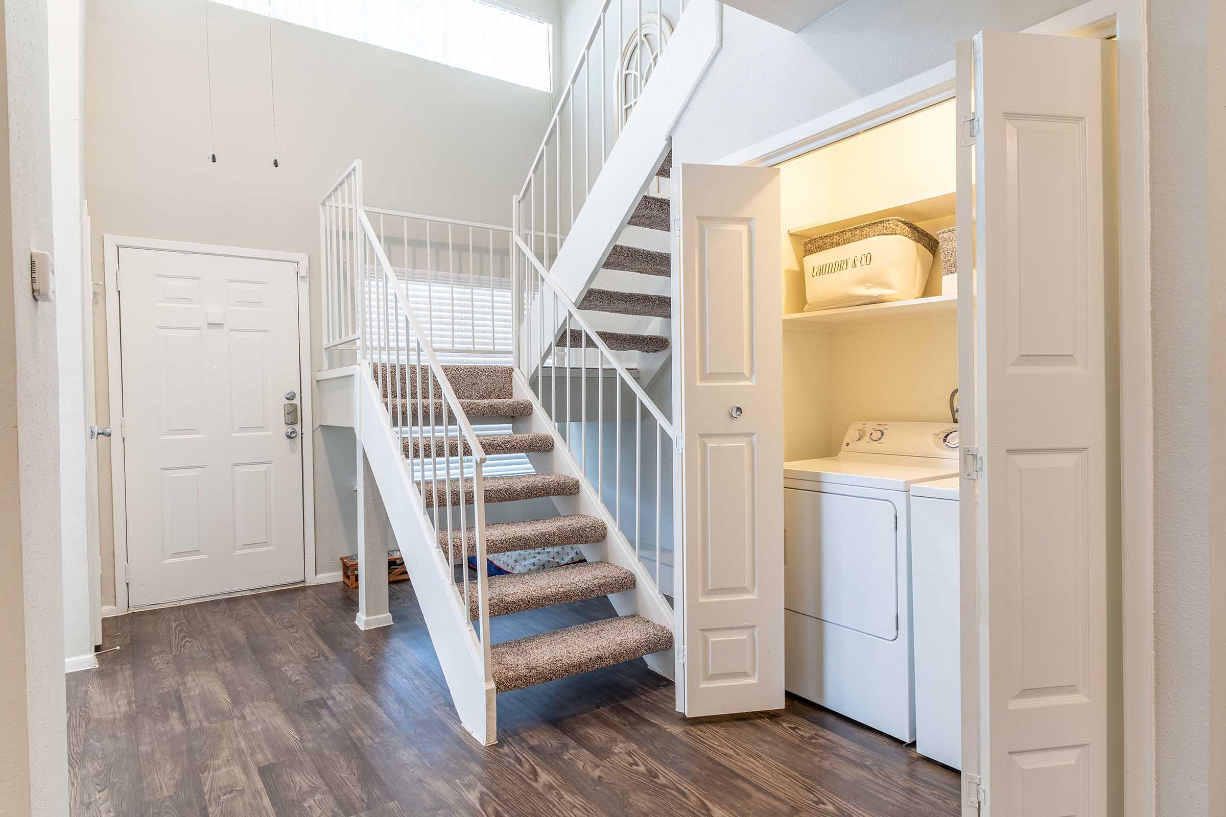 A well-lit entryway featuring a staircase leading to the upper floor. On the right, there is a laundry area with a washer and dryer behind double doors. The floor is covered with wood-style laminate, and the walls are painted in light colors, creating a clean and inviting atmosphere.
