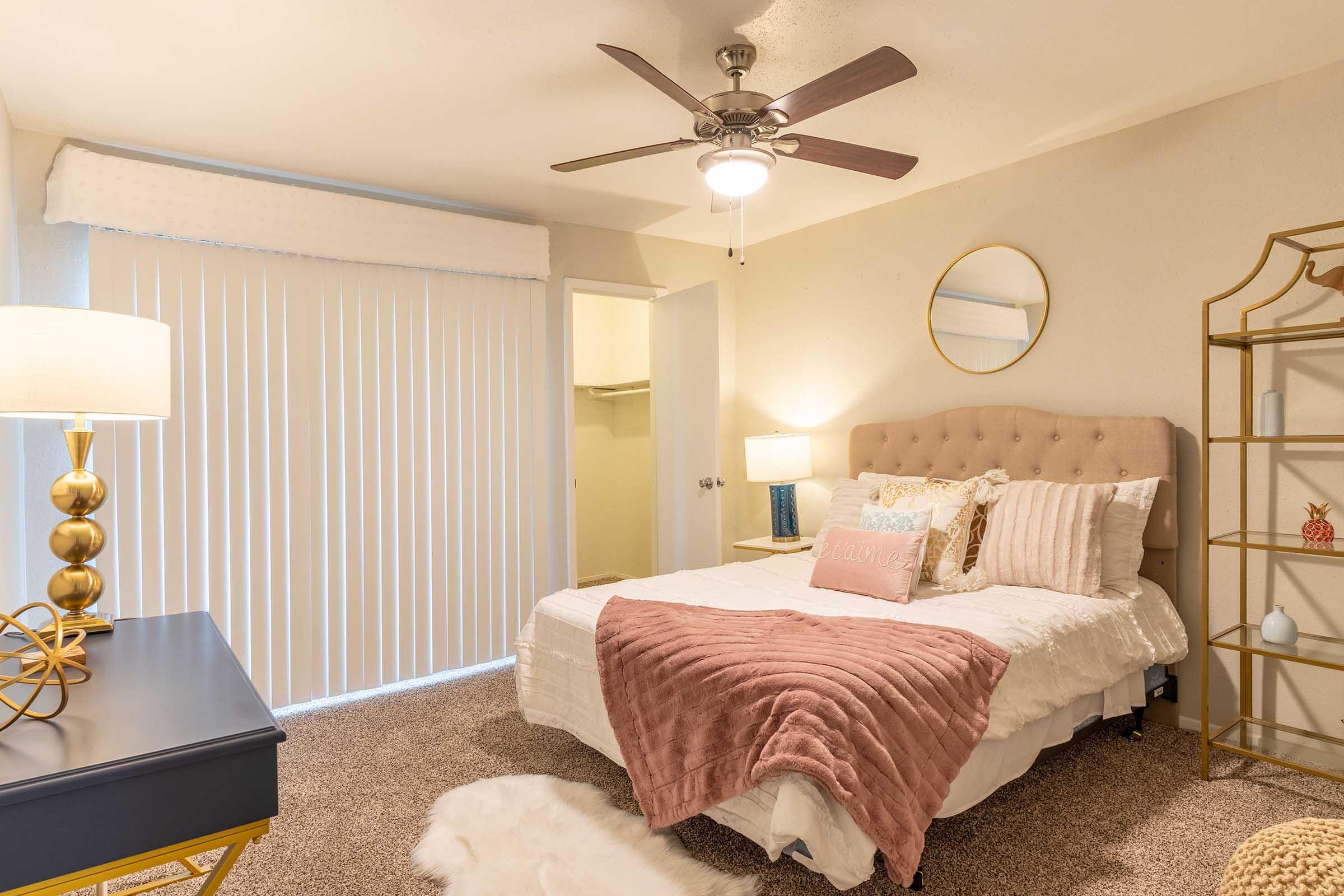 A cozy bedroom featuring a neatly made bed with soft pillows and a decorative throw blanket. There are two lamps on nightstands, a round mirror on the wall, a ceiling fan, and a bookshelf with decorative items. Light-colored walls and a subtle carpet add warmth to the space.