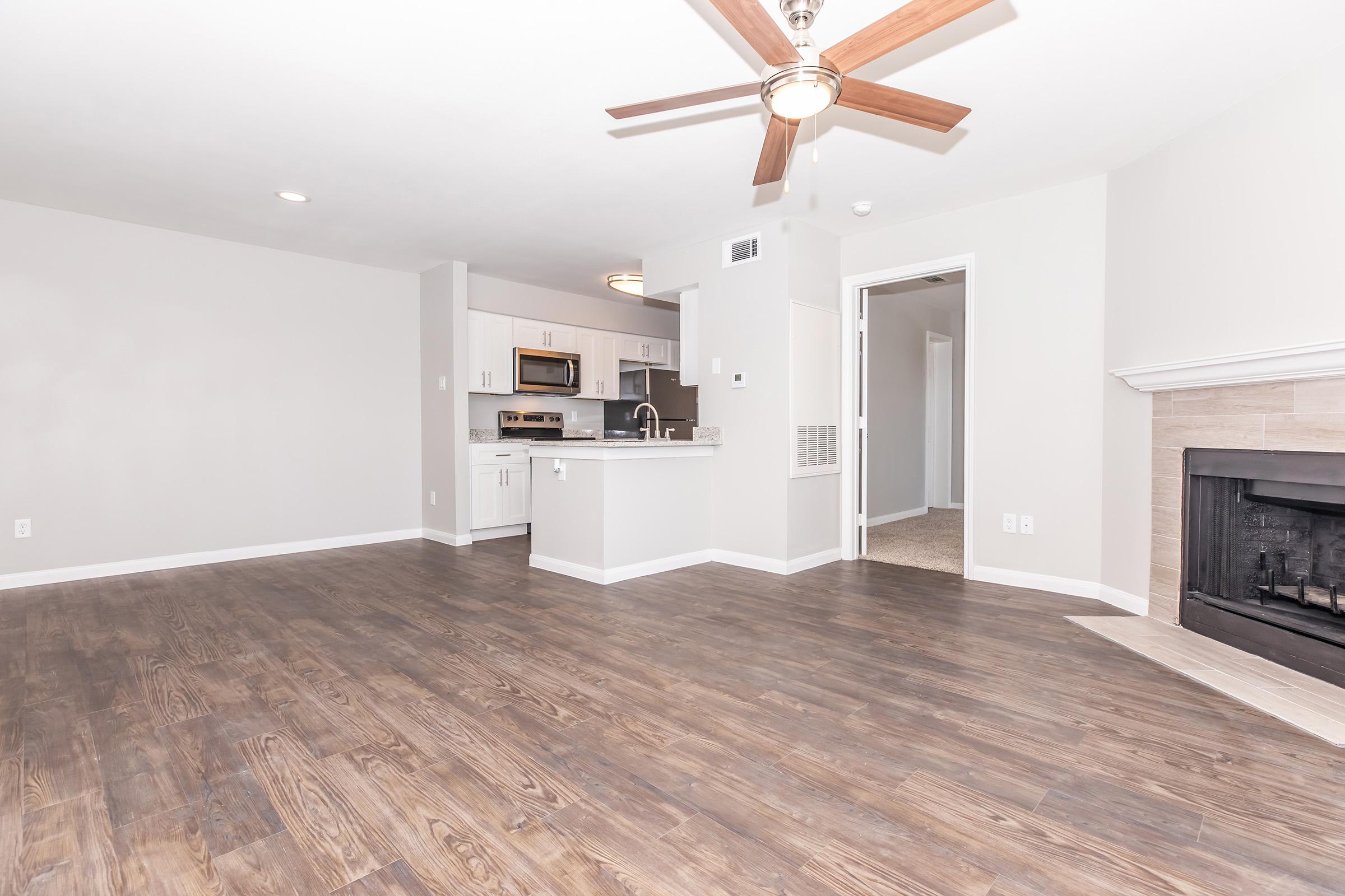 Modern living room interior featuring a ceiling fan, hardwood flooring, and a gas fireplace. The space includes a kitchen with stainless steel appliances and an open layout, leading to a hallway with carpeted flooring. Natural light brightens the area, creating a welcoming atmosphere.