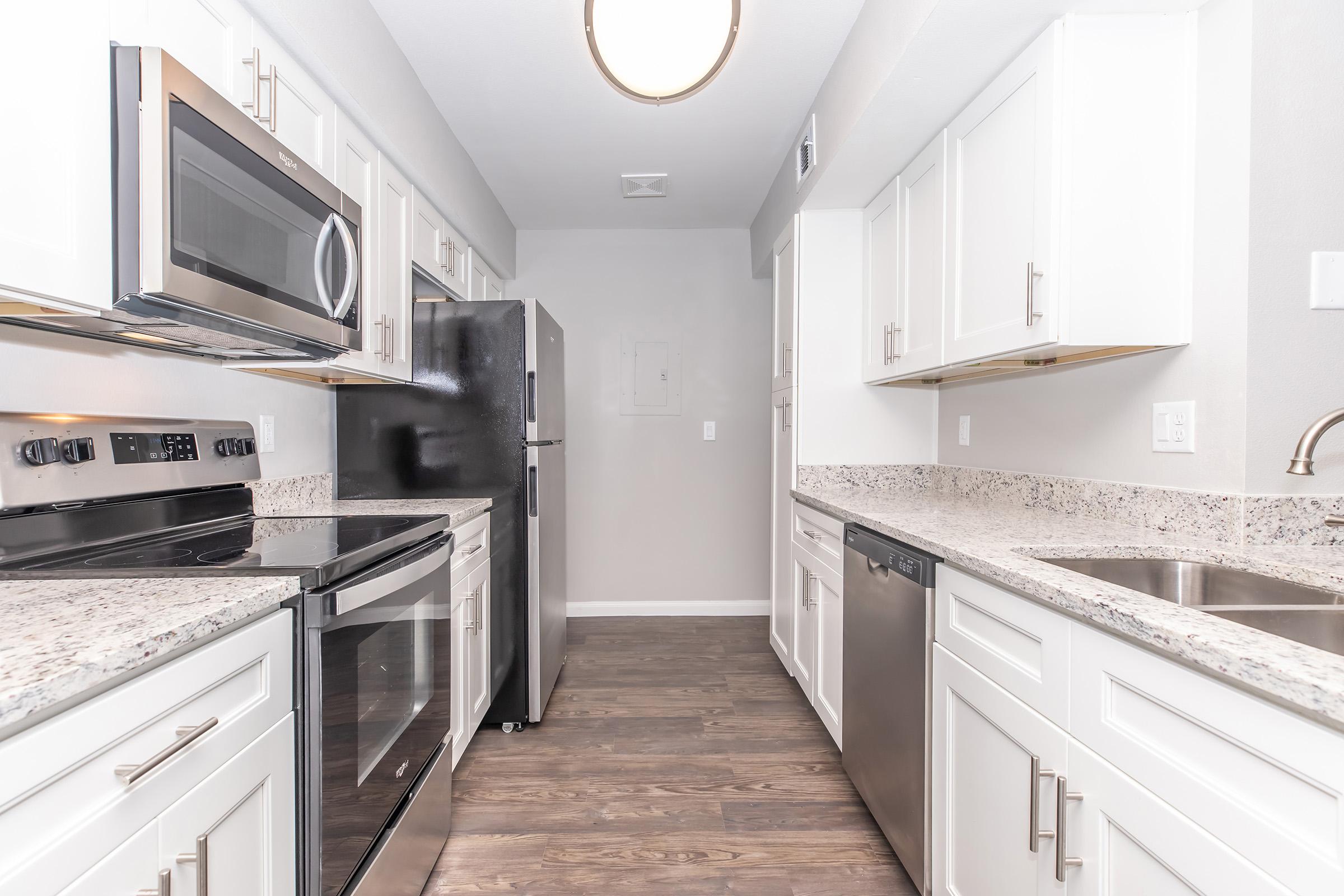Modern kitchen with stainless steel appliances, including a microwave, oven, and refrigerator. The cabinets are white with a glossy finish, and the countertops feature a speckled granite design. There is a double sink and a dishwasher, with wood-style flooring throughout. Bright lighting enhances the spacious feel of the area.