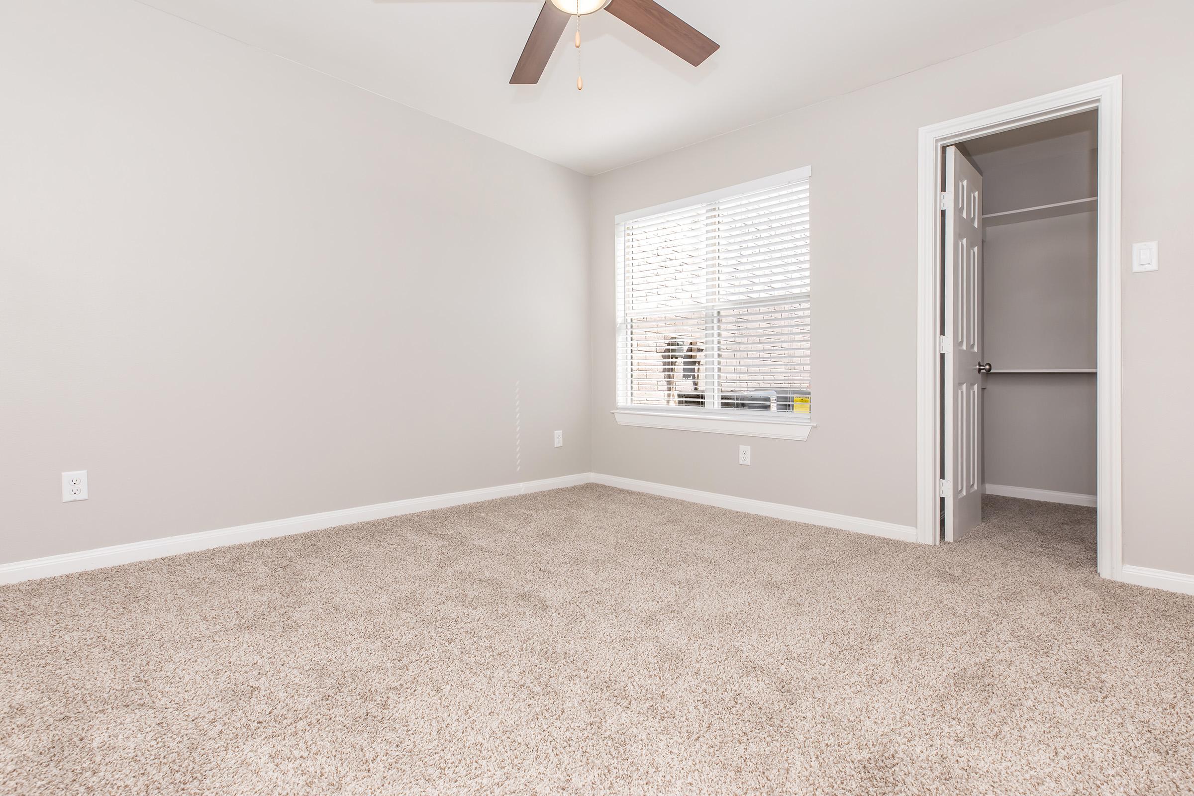 A vacant room with light gray walls and beige carpet. It features a ceiling fan and a window with white blinds allowing natural light. A closet door is visible on the right side, indicating storage space. The overall ambiance is clean and neutral, suitable for various purposes.