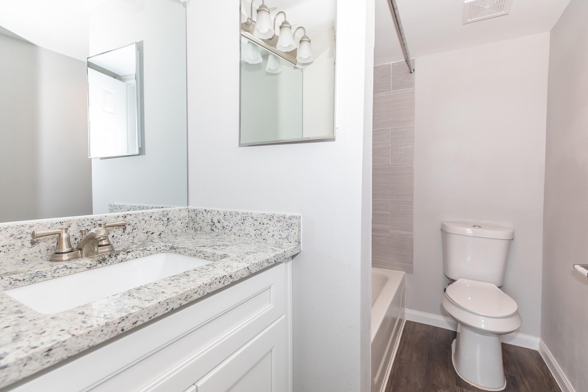 A modern bathroom featuring a white vanity with a granite countertop, a mirror above the sink, and a well-lit area. There is a bathtub with a shower curtain, a toilet, and neutral-colored walls and flooring, creating a clean and spacious look.