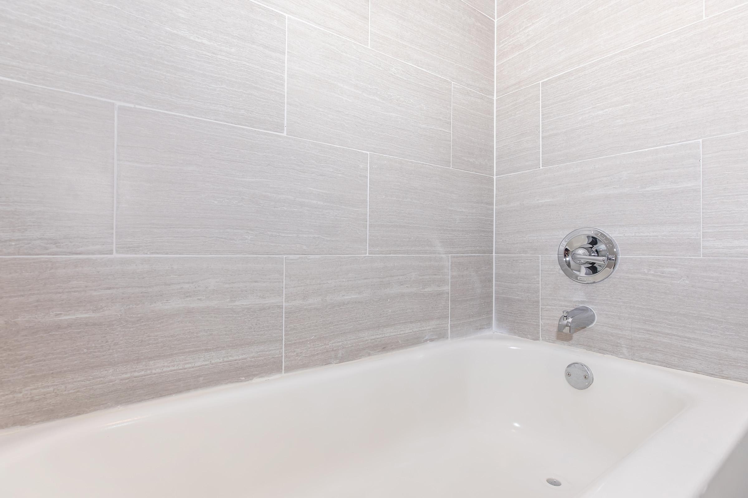 A modern bathroom featuring a clean, empty bathtub with a smooth, white surface. The walls are covered in light gray tiles, creating a minimalist aesthetic. The faucet and shower control are visible on the wall, complementing the contemporary design. Natural light enhances the spacious feel.
