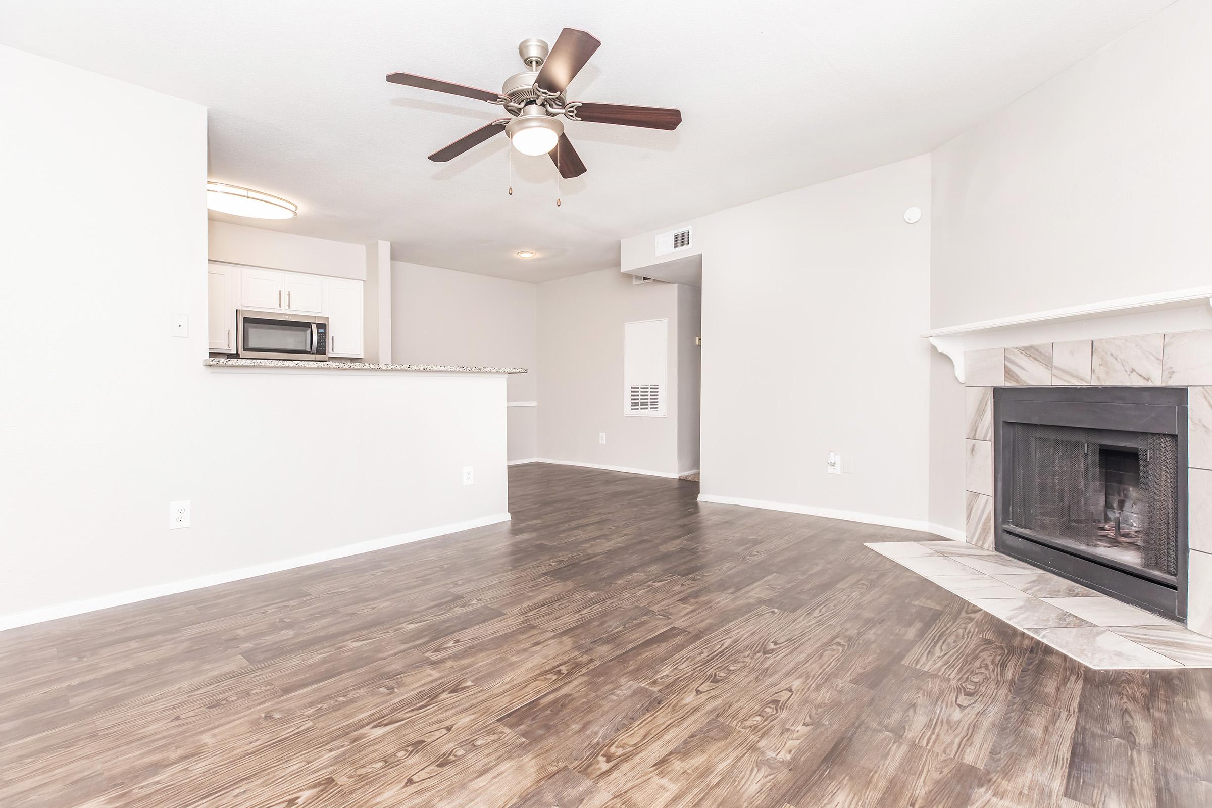 Spacious living area featuring a ceiling fan, hardwood-style flooring, and a cozy fireplace. The open layout includes a kitchen area with modern appliances visible in the background. Light, neutral wall colors enhance the bright and airy atmosphere of the space.
