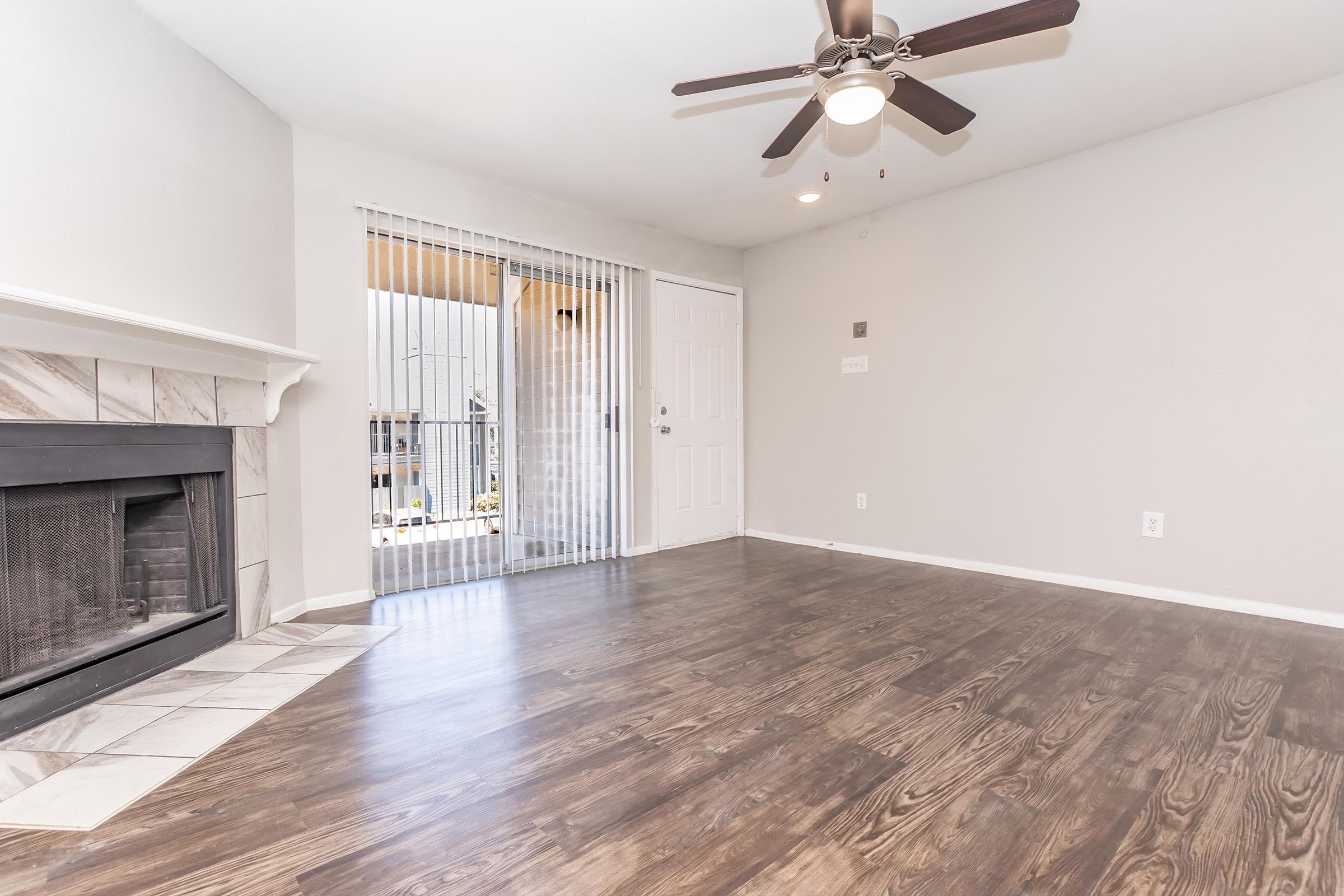 Interior view of a spacious, well-lit living room featuring a fireplace with a tile surround, large windows with vertical blinds, and a ceiling fan. The floor is adorned with dark wood laminate, complementing the neutral-colored walls. A door leads outside, enhancing the room's open feel.