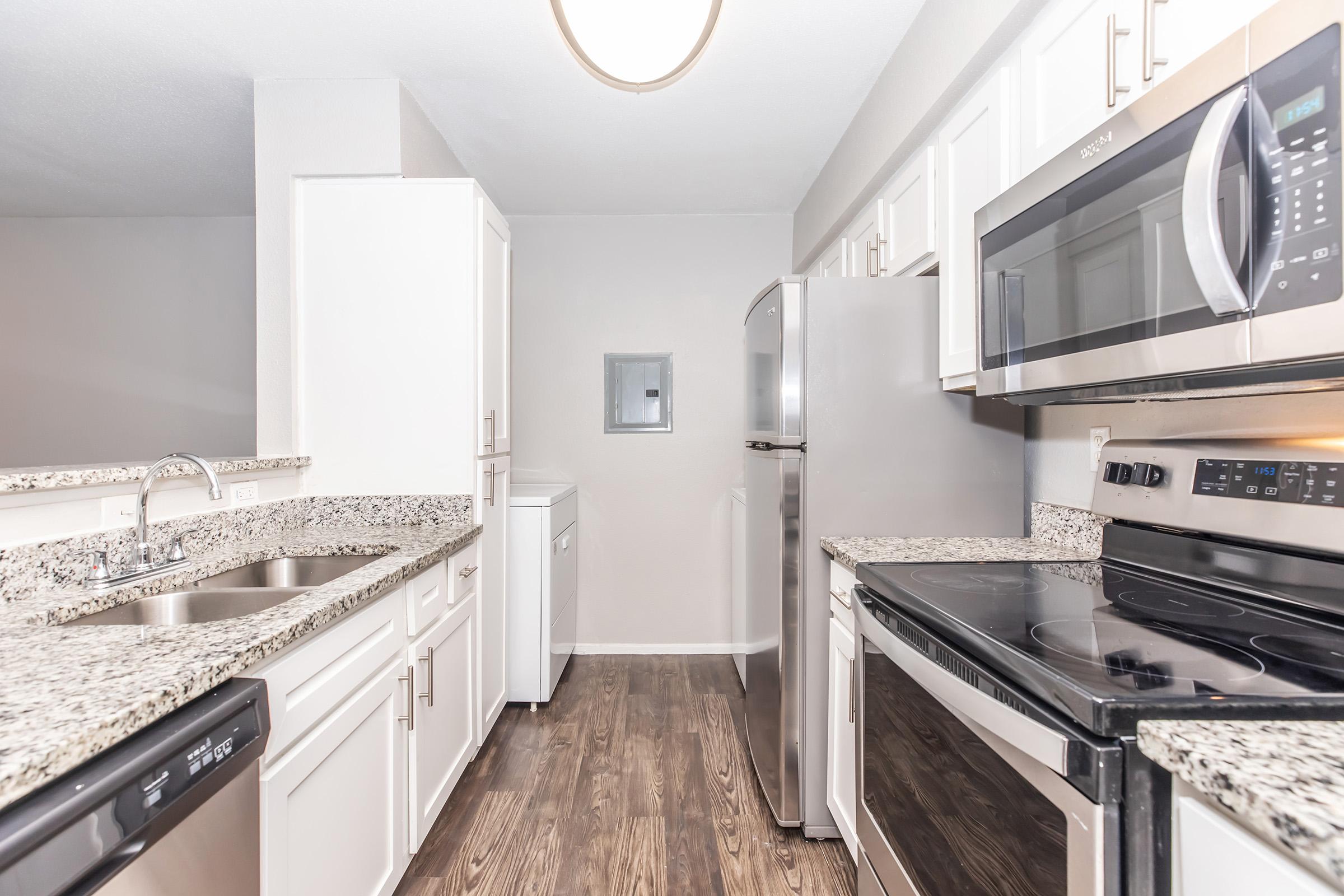 Modern kitchen featuring white cabinets, granite countertops, stainless steel appliances, including a refrigerator, microwave, and oven. The space has an open layout, with a view of the dining area. The flooring is a warm, wood-like finish, contributing to a cozy atmosphere. Ample natural light enhances the airy feel.