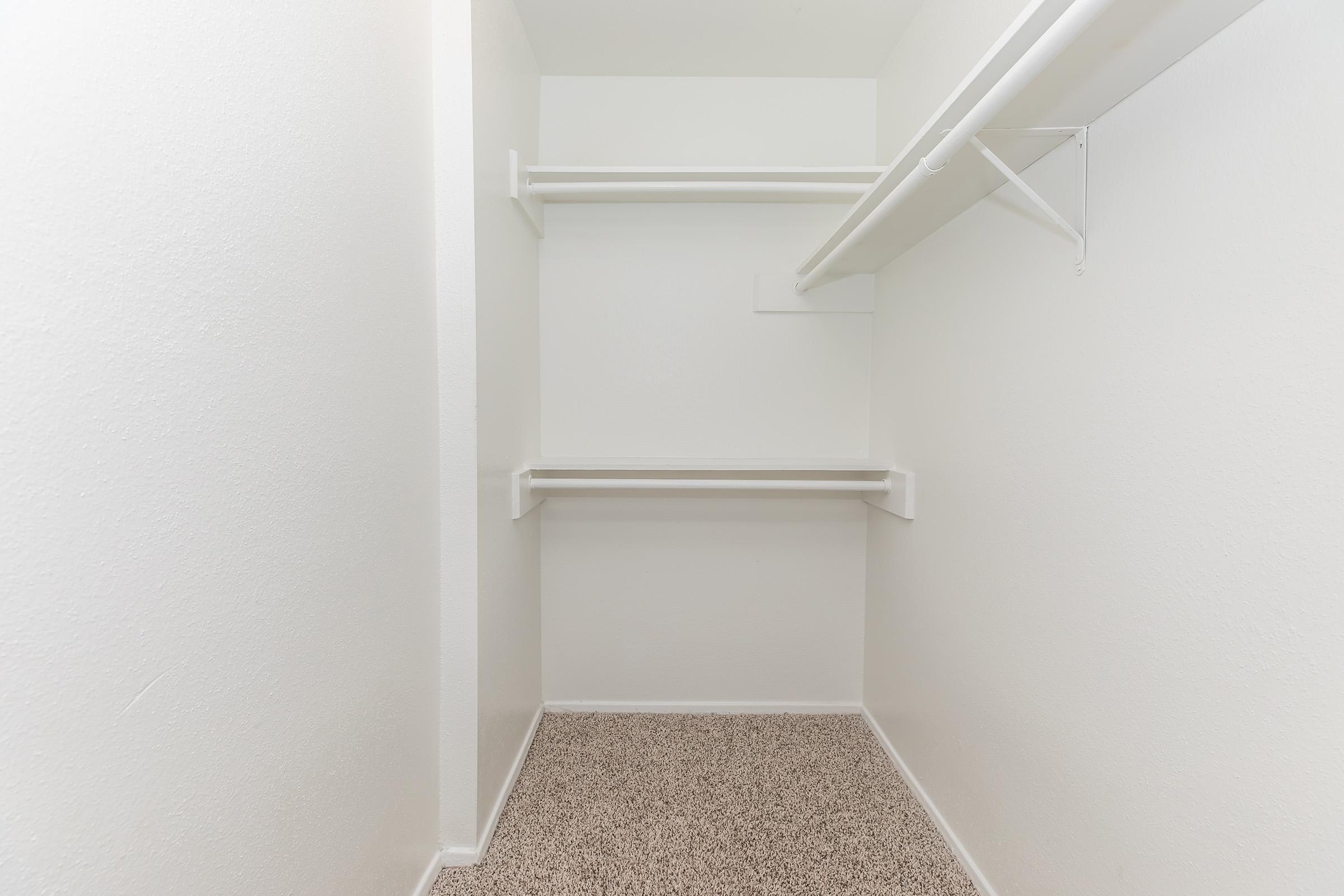 Empty closet with white walls, featuring two hanging rods and a single shelf. The floor is covered with light-colored carpet. The space is well-lit and organized, creating a clean and minimalist look.