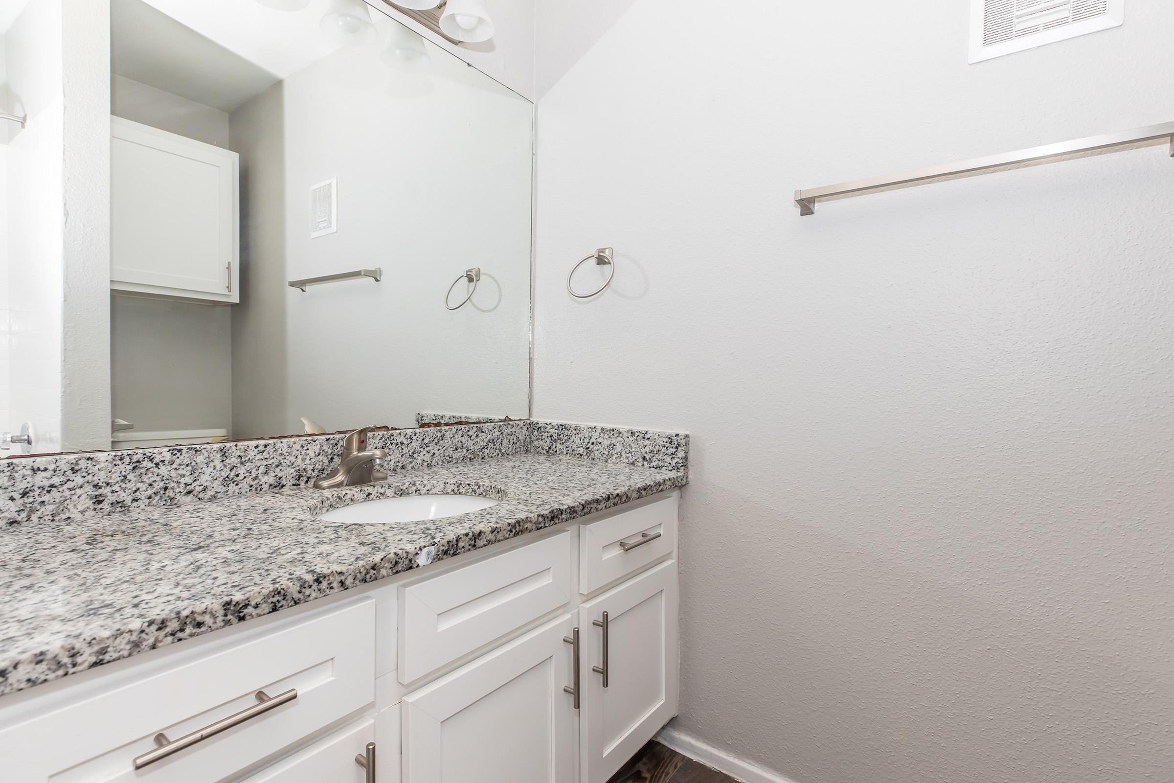 A modern bathroom featuring a gray wall, a white cabinetry with a granite countertop, a sink, and a large mirror. The space is well-lit, with a towel bar and minimal decor, creating a clean and spacious appearance.