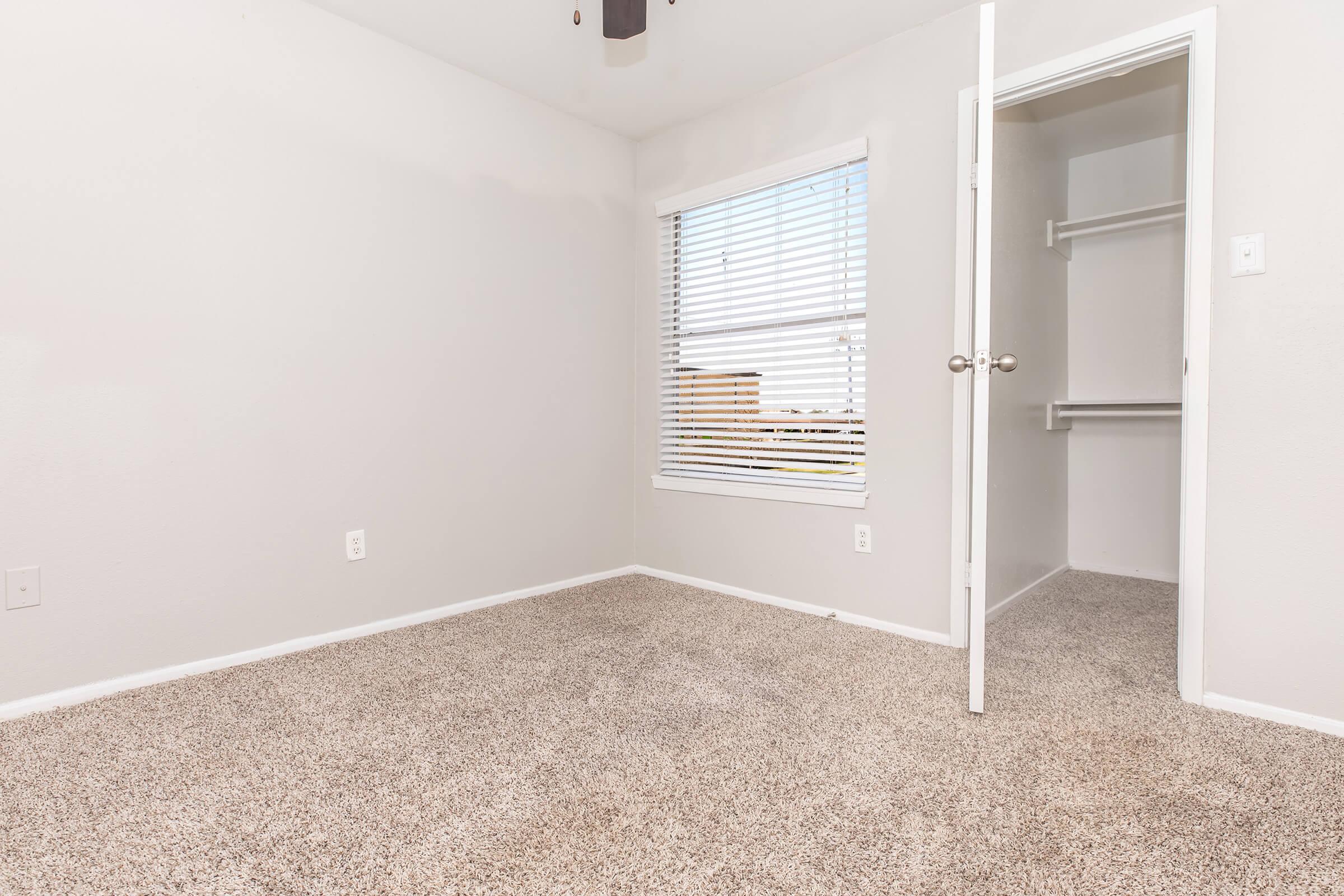 A small, empty room with light gray walls and carpet. There's a window with white blinds allowing natural light to enter. A closet is visible through an open door on the right, featuring a simple rod for hanging clothes. The space appears clean and ready for furnishings.