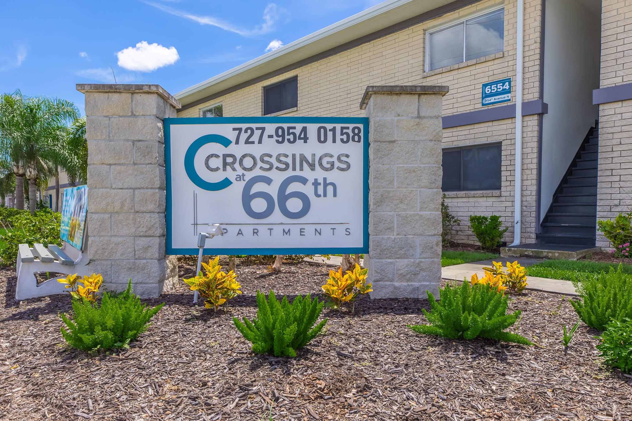 Sign for "Crossings at 66th Apartments" with contact number 727-954-0158. Surrounding landscape features well-maintained plants and a small bench, against a backdrop of a multi-unit building. Bright blue sky and clouds are visible above.