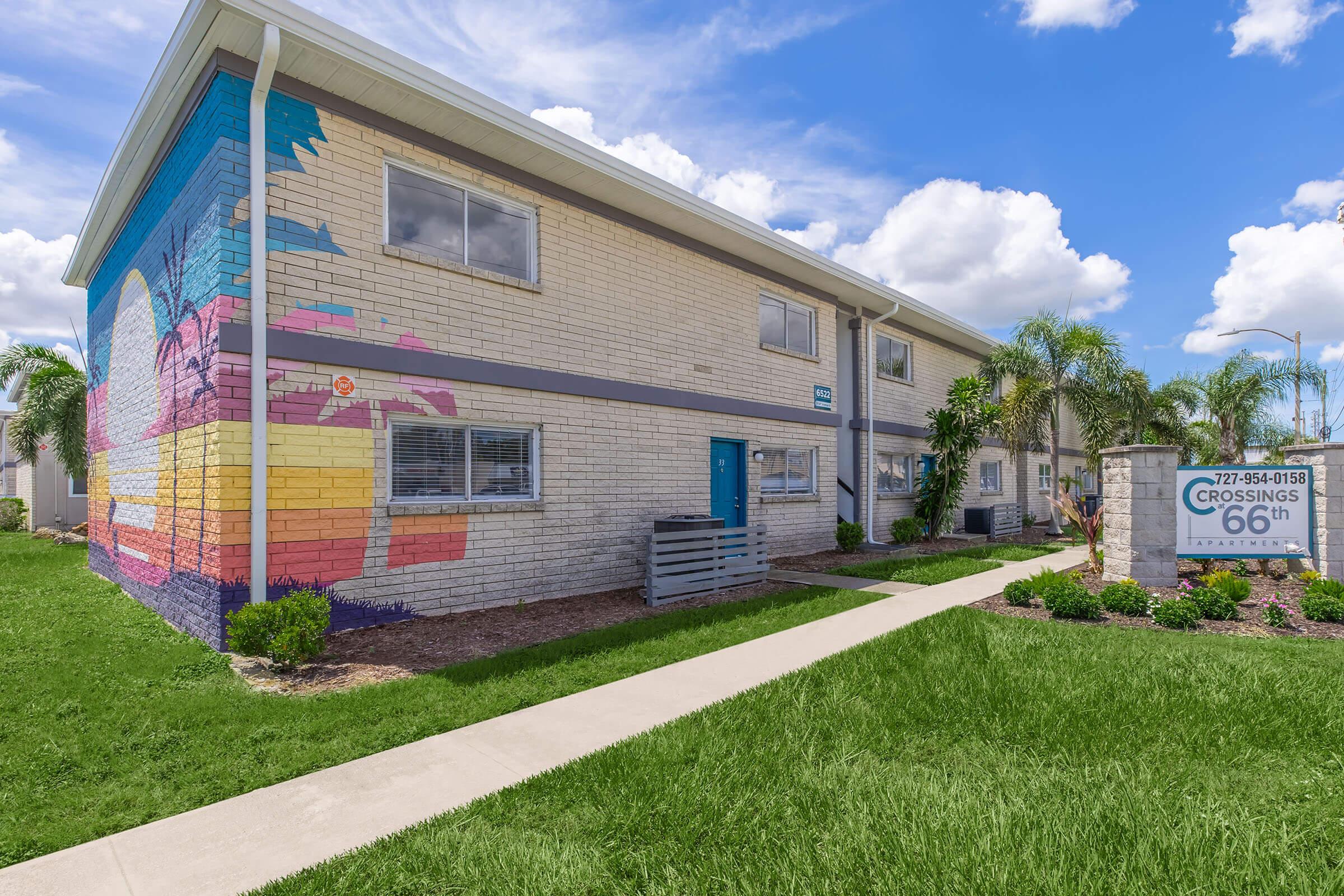 A two-story apartment building with a colorful mural featuring palm trees and a sunset on one side. The property features a well-maintained lawn with green grass and palm trees. A sign in front displays "Crossings 66th" along with a contact number. The sky is bright blue with white clouds.