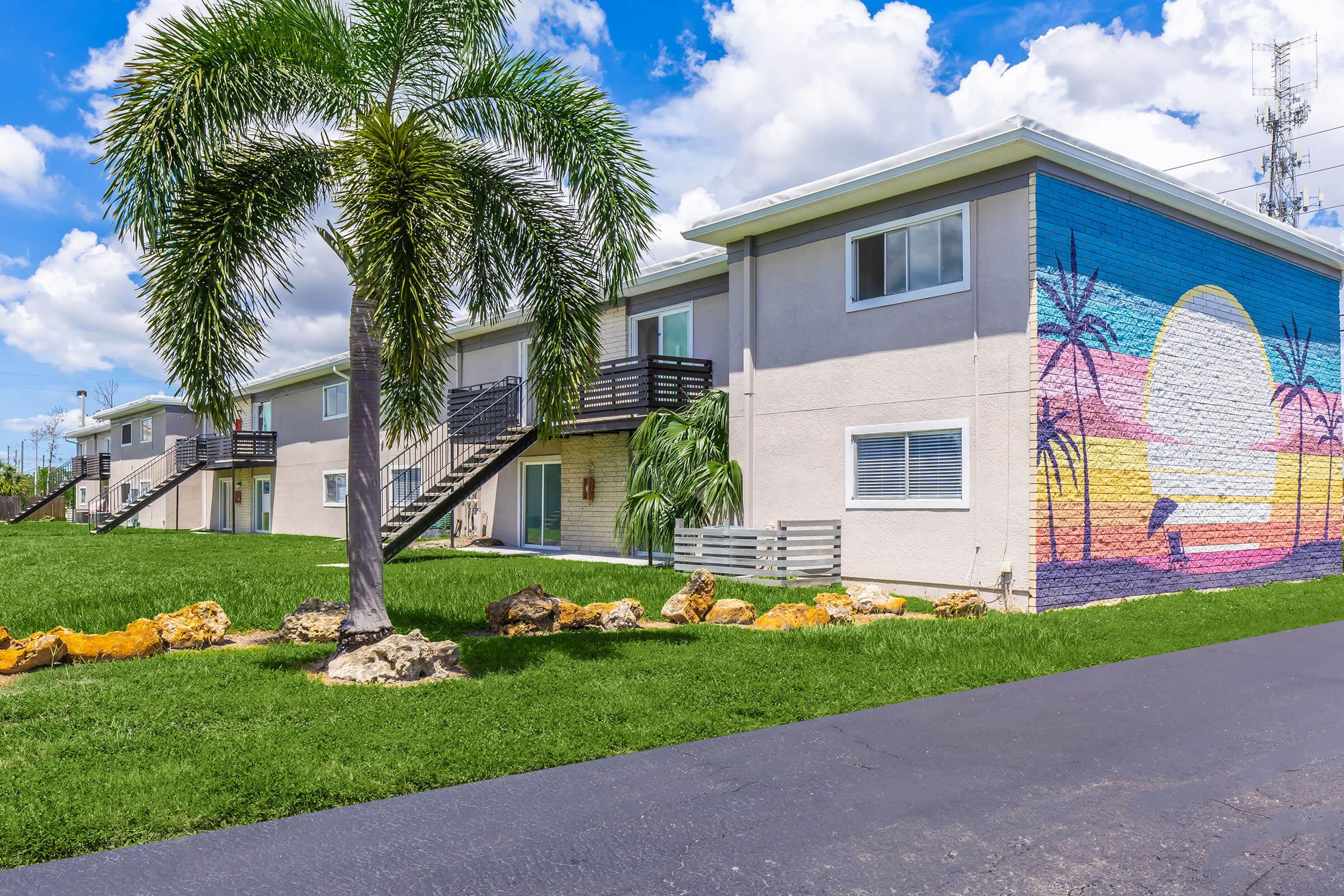 A colorful mural depicting a sunset with palm trees on the side of a building. The building features two stories, with balconies and a grassy area in front. Lush palm trees and rocks are part of the landscaping, under a bright blue sky with fluffy white clouds.