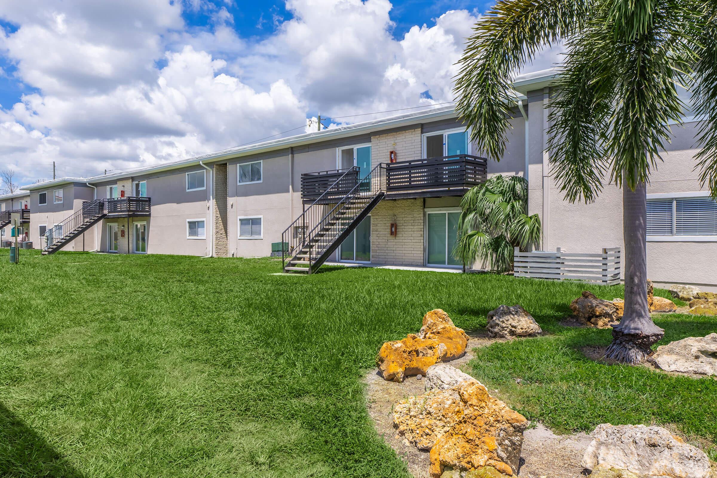 Exterior view of a multi-unit apartment building with two levels. The building features balconies with stairs leading down to a grassy area. Lush green landscaping includes a palm tree and decorative stones. The sky is partly cloudy, creating a bright and inviting atmosphere.