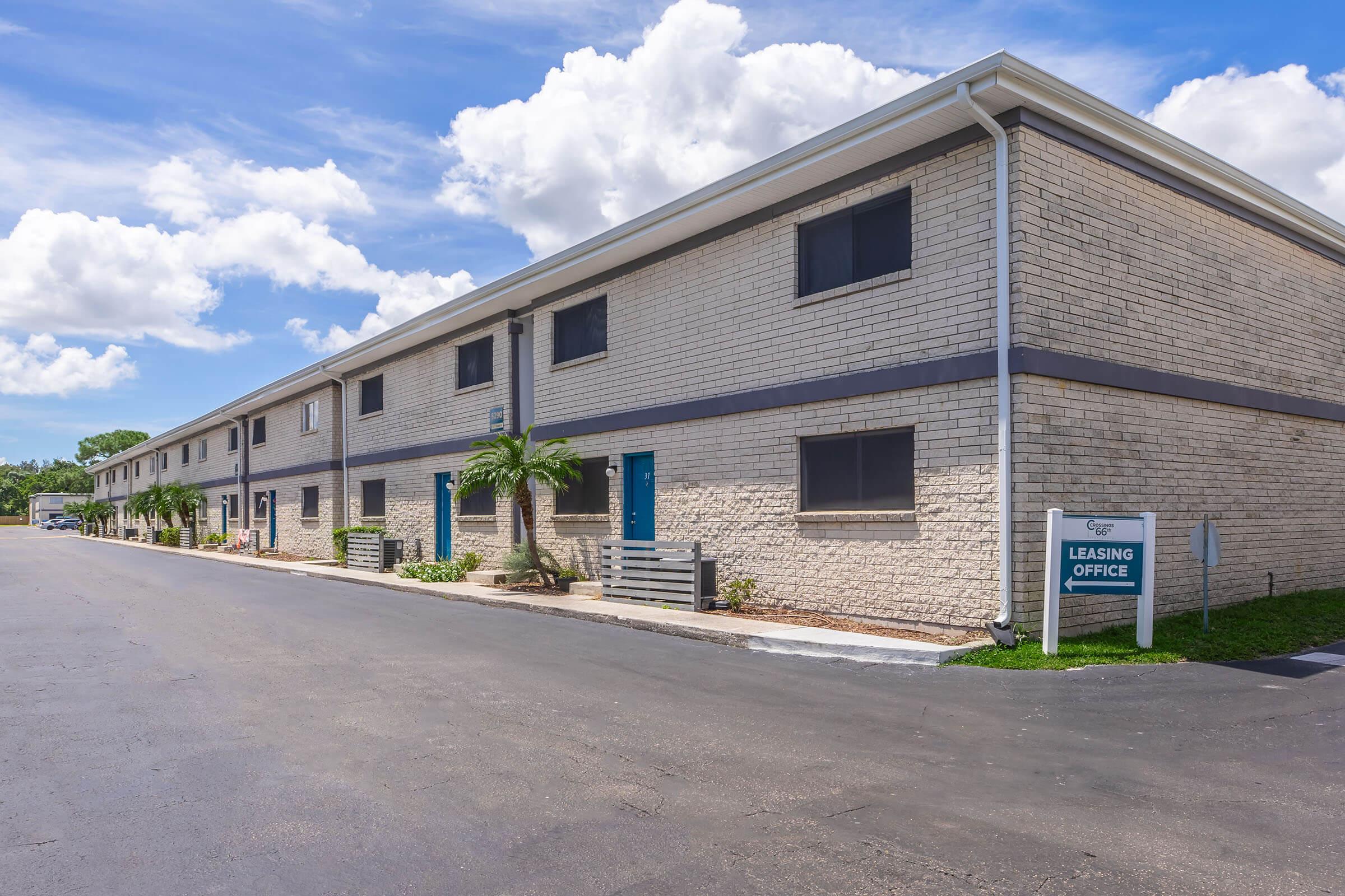 A row of residential buildings with a leasing office in view. The structure features beige brick walls, blue doors, and small palm trees planted along the walkway. The sky is partly cloudy, and a paved road runs in front of the buildings. A sign indicates the location of the leasing office.