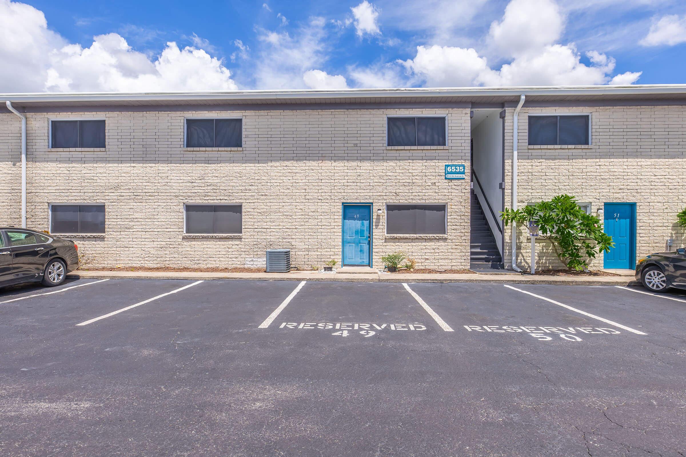 A multi-story building with a light-colored brick exterior, featuring blue doors. The front has designated parking spaces labeled "RESERVED." Lush greenery is visible near the entrance, and the sky is partly cloudy, adding a vibrant contrast to the scene.