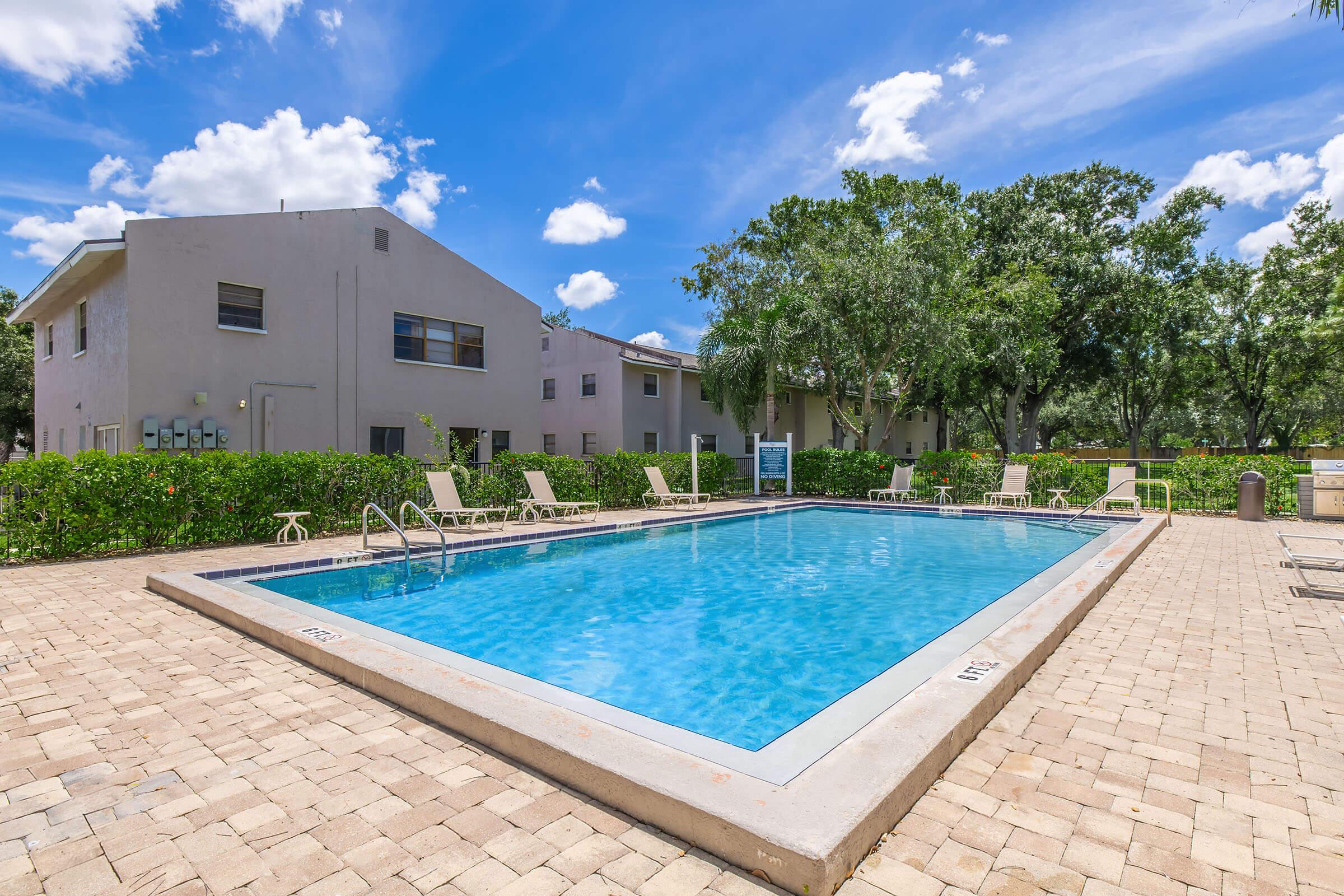 A sunny outdoor swimming pool surrounded by lush greenery and residential buildings. The pool area features lounge chairs and a clear blue sky with scattered clouds. The deck is paved with light-colored stones, creating a relaxing atmosphere for leisure and recreation.