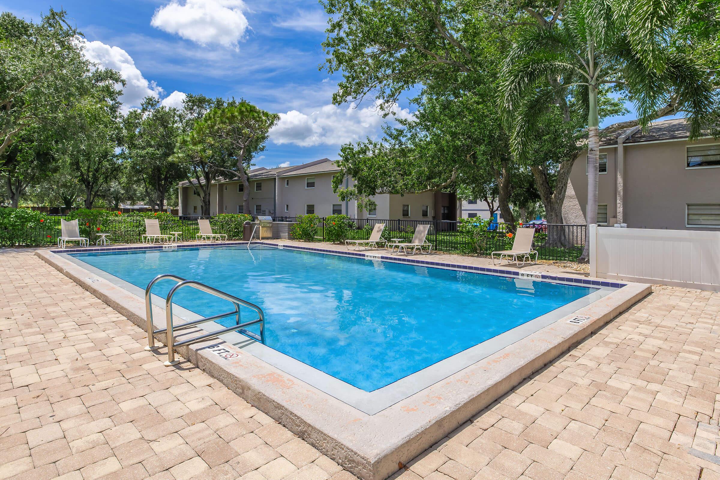 A bright blue swimming pool surrounded by sun loungers on a stone patio. Lush greenery and trees are in the background, with residential buildings partially visible. The sky is clear with a few fluffy clouds, creating a scenic outdoor setting perfect for relaxation.