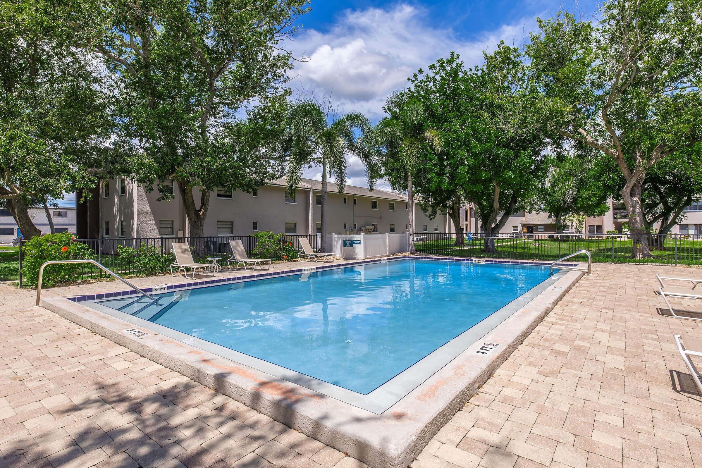 A clear outdoor swimming pool surrounded by tropical trees and manicured lawns. Lounge chairs are positioned around the pool area, with nearby residential buildings visible in the background under a partly cloudy sky. The scene conveys a tranquil, inviting atmosphere perfect for relaxation.