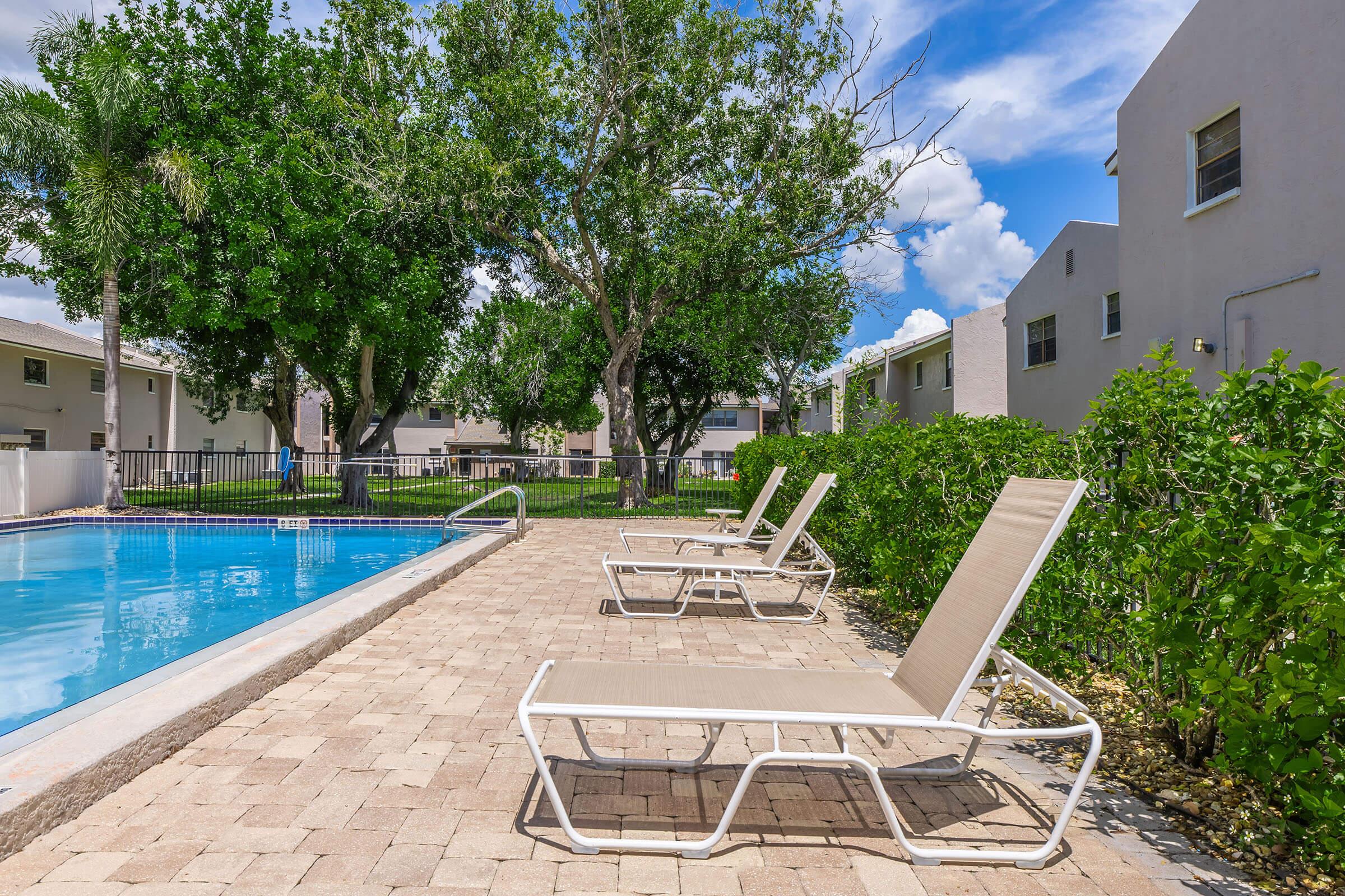 A scenic view of a swimming pool surrounded by green trees and manicured shrubs. White lounge chairs are lined up beside the pool on a stone patio, with a clear blue sky and fluffy clouds in the background. Nearby buildings are visible, adding to the tranquil atmosphere.