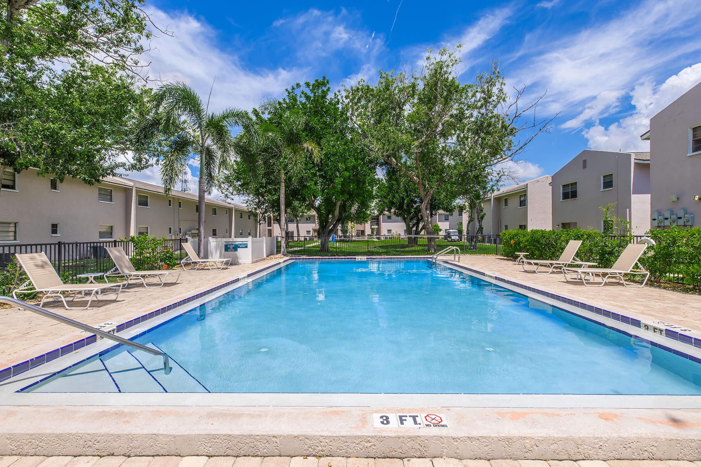 A clear blue swimming pool surrounded by lounge chairs and a well-maintained lawn. Lush green trees provide shade, and residential buildings are visible in the background under a bright blue sky with scattered clouds. The pool depth indicator shows "3 FT" at one end.