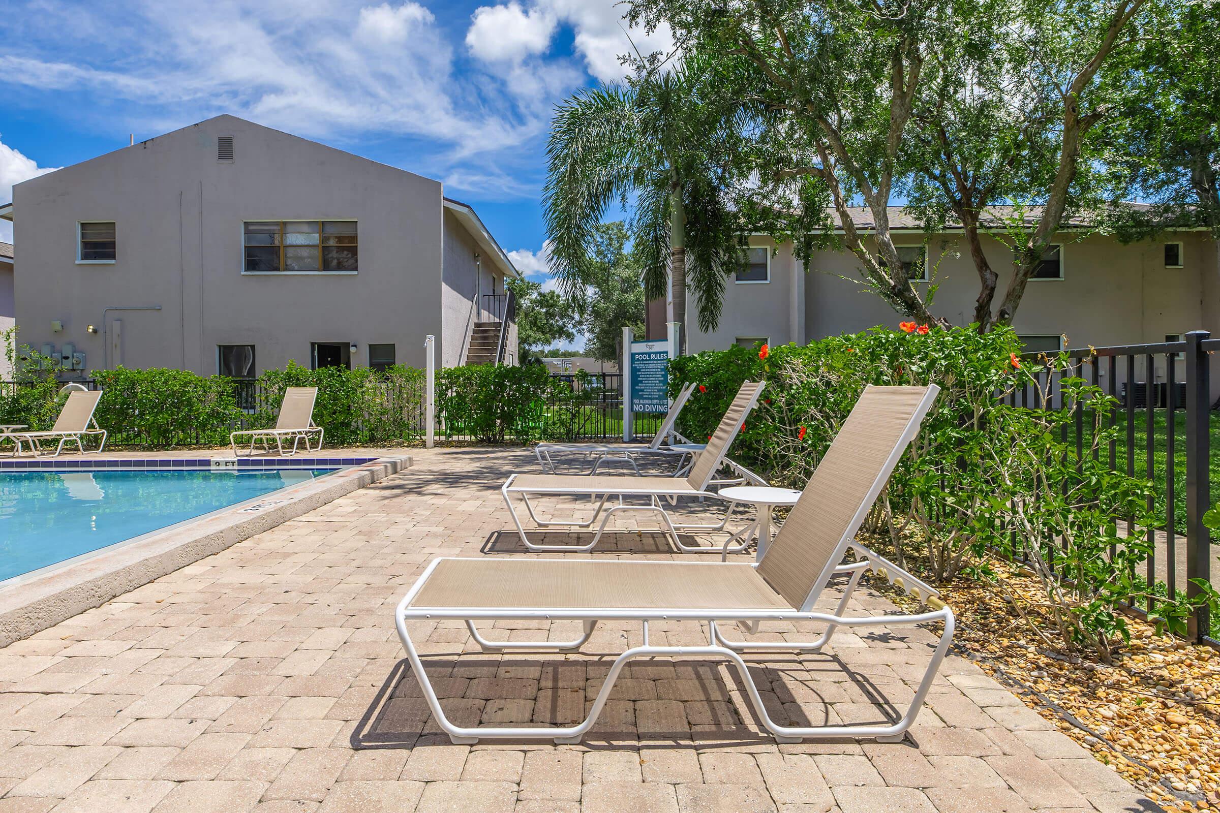 A sunny pool area featuring lounge chairs arranged around a swimming pool. In the background, there are two apartment buildings surrounded by lush greenery and shrubs. Clear blue skies with fluffy white clouds overhead. The scene conveys a relaxed, inviting atmosphere for residents and guests.
