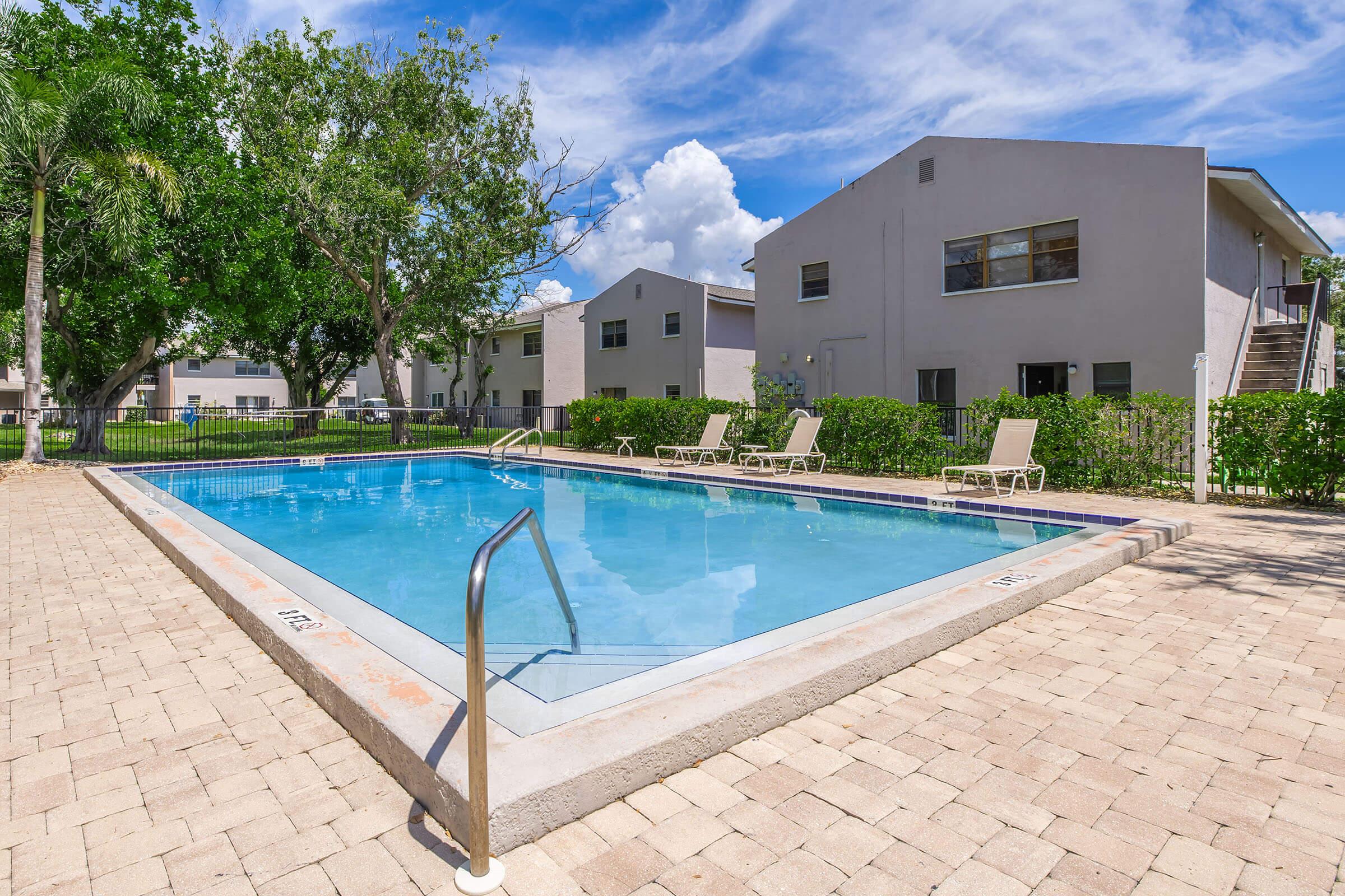 A clear swimming pool surrounded by lounge chairs, set within a well-maintained area with green shrubs and trees. In the background, modern residential buildings are visible under a blue sky with scattered clouds.