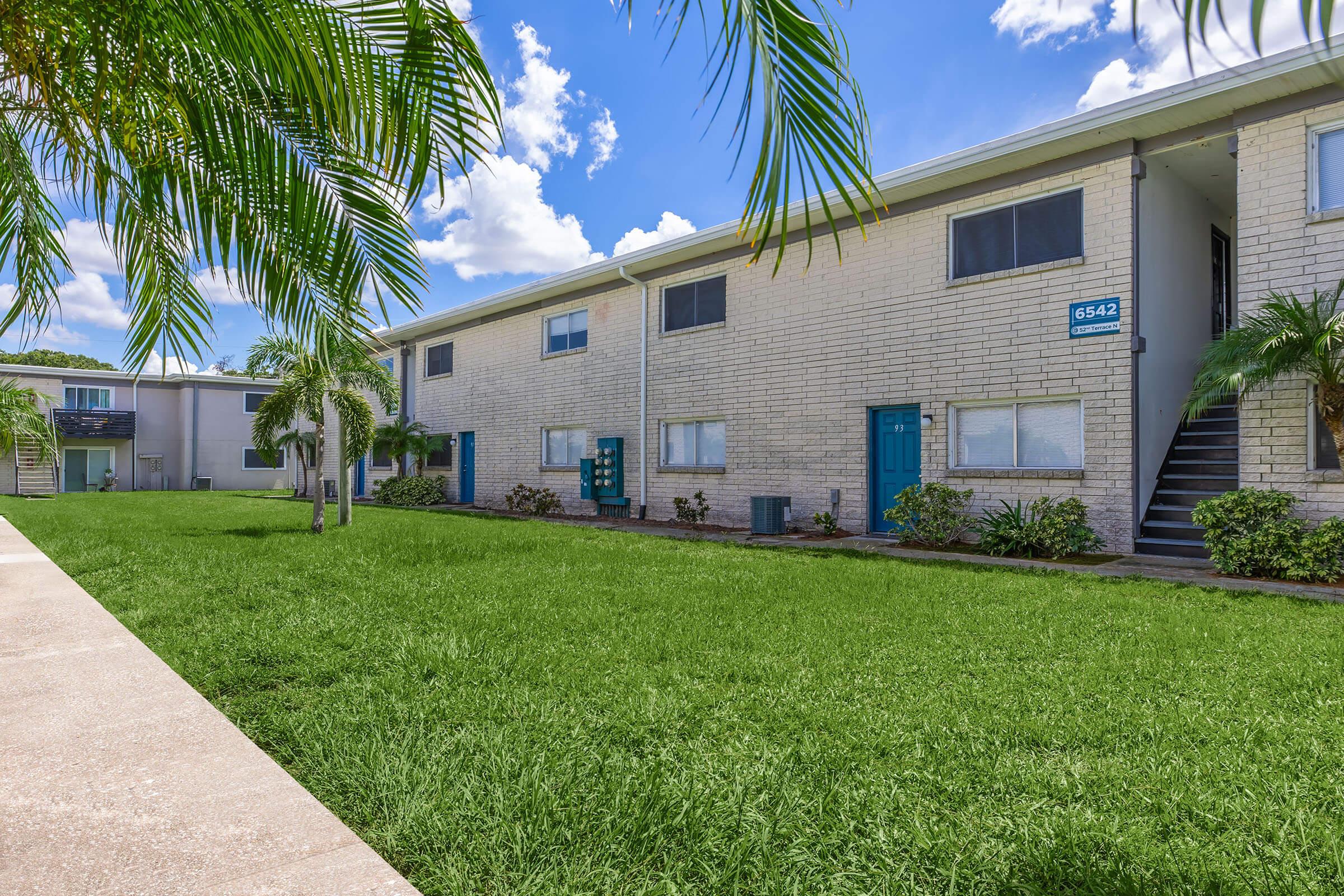 A well-maintained apartment complex featuring beige brick buildings with blue doors, surrounded by lush green lawns and palm trees. The photo captures a bright, sunny day with blue skies and scattered clouds, enhancing the inviting atmosphere of the residential area.