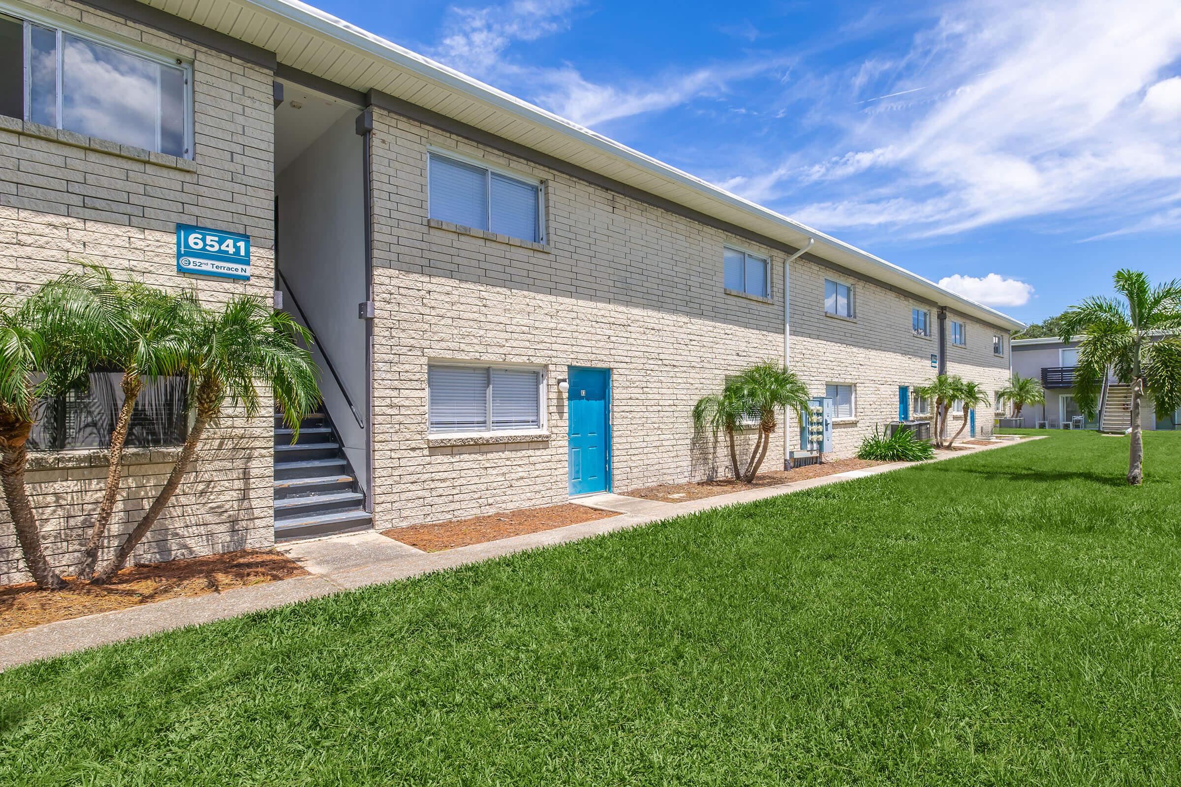 A modern apartment building with a light brick exterior and blue doors. There are palm trees and well-maintained green grass in the foreground. The sky is bright blue with a few fluffy clouds, creating a welcoming atmosphere. The scene showcases a tidy and inviting living space.
