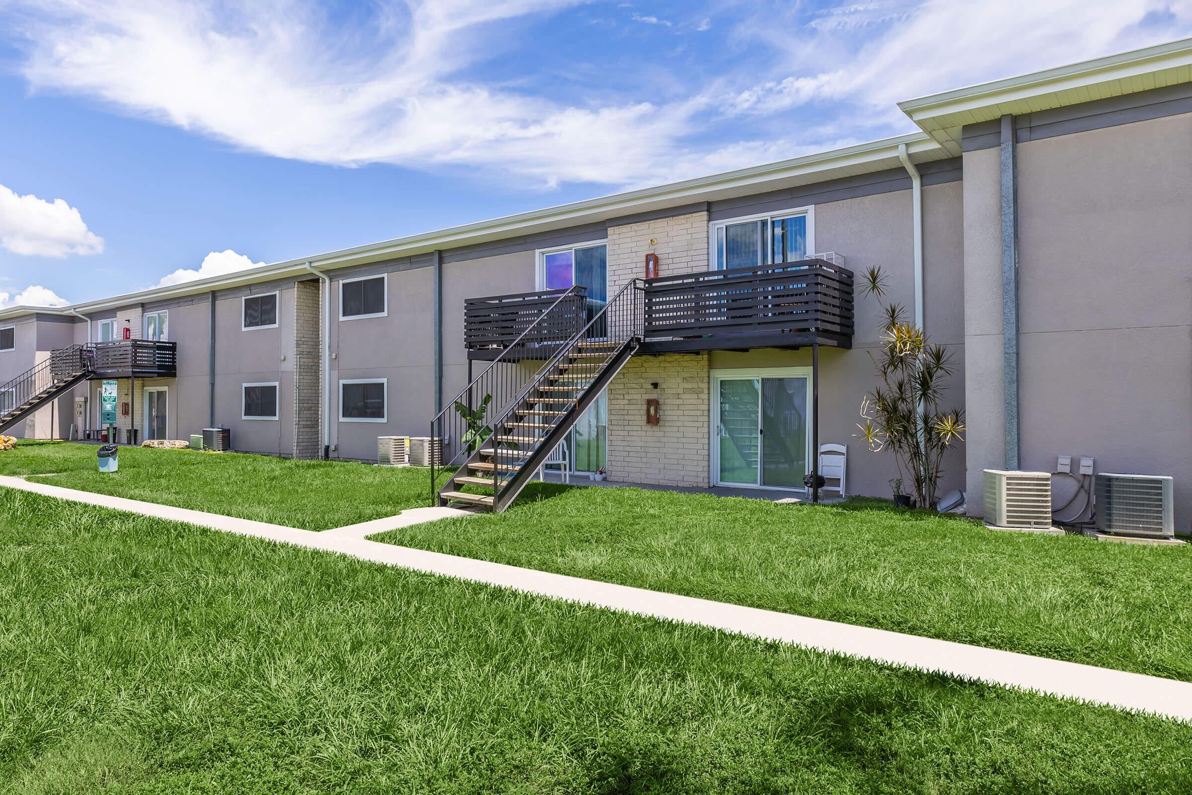 View of a modern apartment complex featuring two-story buildings with balconies, green grass in the foreground, and a paved walkway leading to the entrances. Air conditioning units are visible on the ground, and decorative plants are seen near the stairs. The sky is partly cloudy, adding a bright atmosphere to the scene.