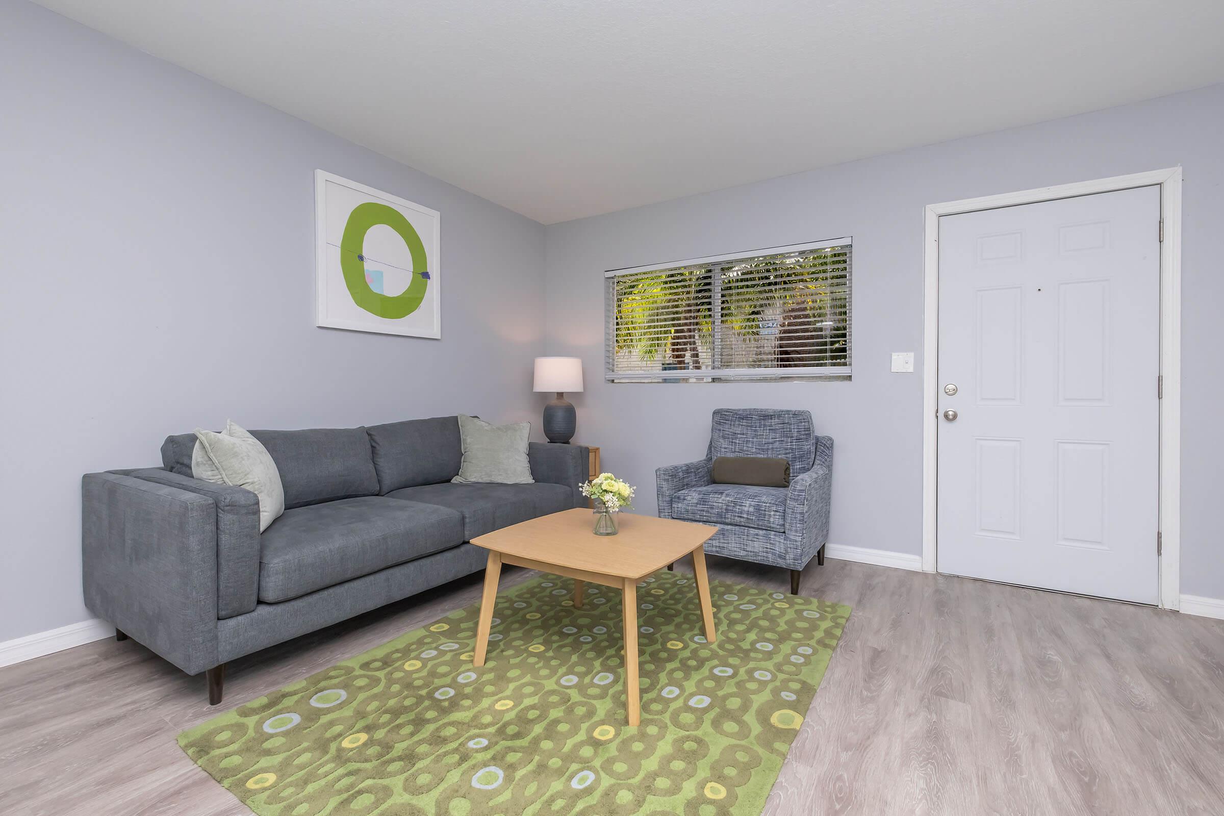 A modern living room featuring a gray sofa with decorative pillows, an armchair in a matching tone, a wooden coffee table with a small vase of yellow flowers, and a colorful area rug. The walls are painted light gray, and there is a window with blinds and a minimalistic wall art piece above the sofa.