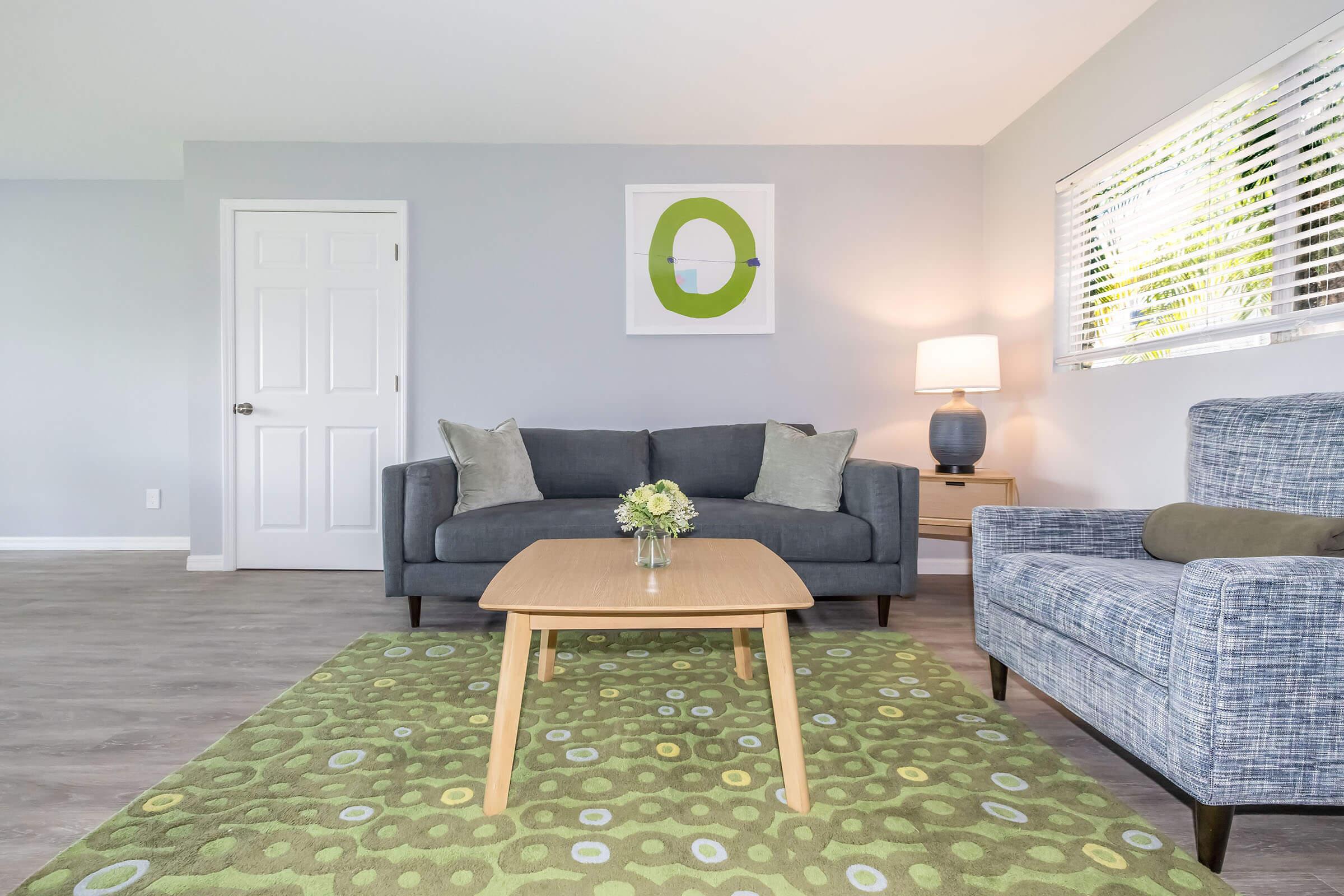 A modern living room featuring a gray sofa with decorative pillows, a light wooden coffee table on a green patterned rug, and a blue armchair. A small vase of flowers sits on the table, while a lamp provides warm lighting. A piece of abstract art adorns the wall, and there's a door on the left.