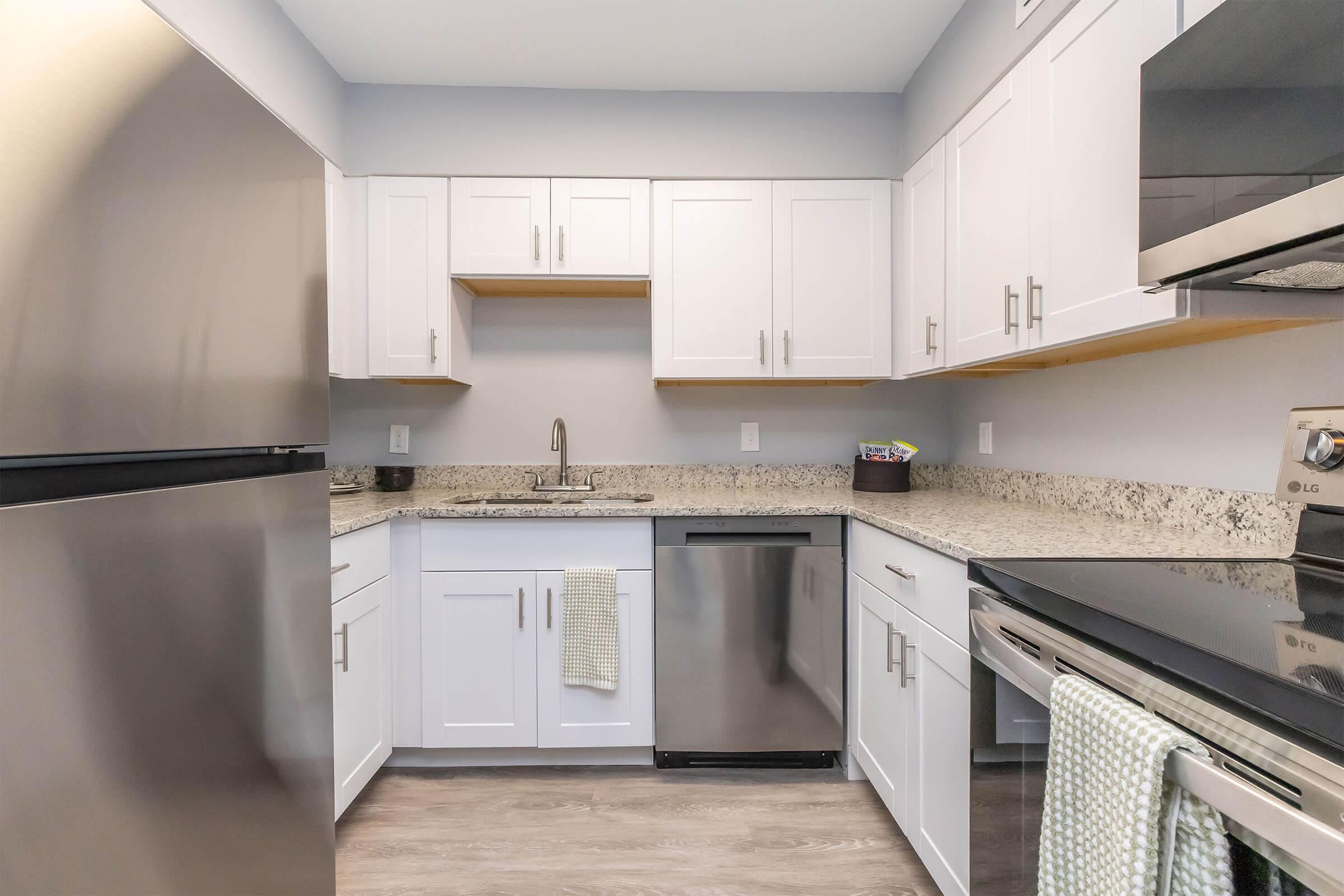 A modern kitchen featuring white cabinets, granite countertops, a stainless steel refrigerator, a dishwasher, and an electric stove. The space is well-lit with a neutral color palette, showcasing a clean and minimalist design. A dish towel hangs from the oven handle, adding a touch of home.
