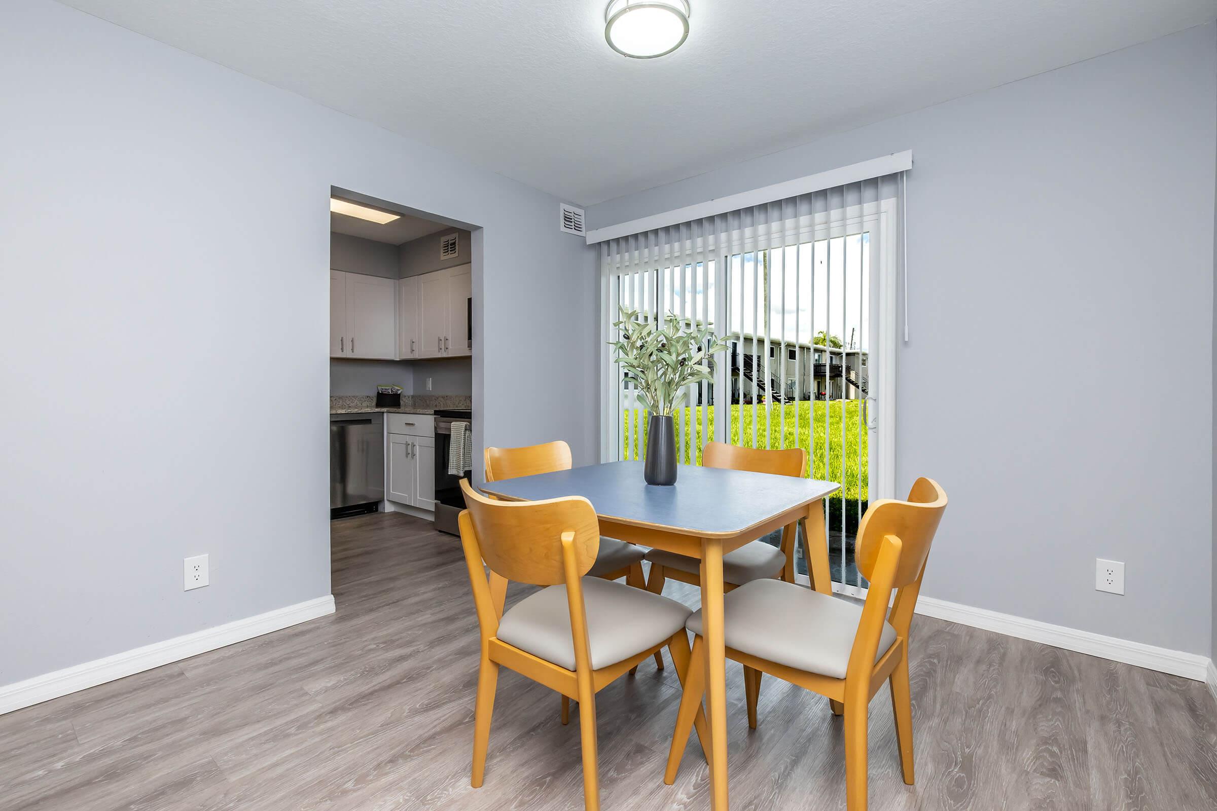 A bright and modern dining area featuring a wooden table with four light-colored chairs. A vase with greenery sits in the center of the table. In the background, there is a sliding glass door leading to an outdoor space, and a kitchen area is partially visible through an archway. The walls are painted light gray.