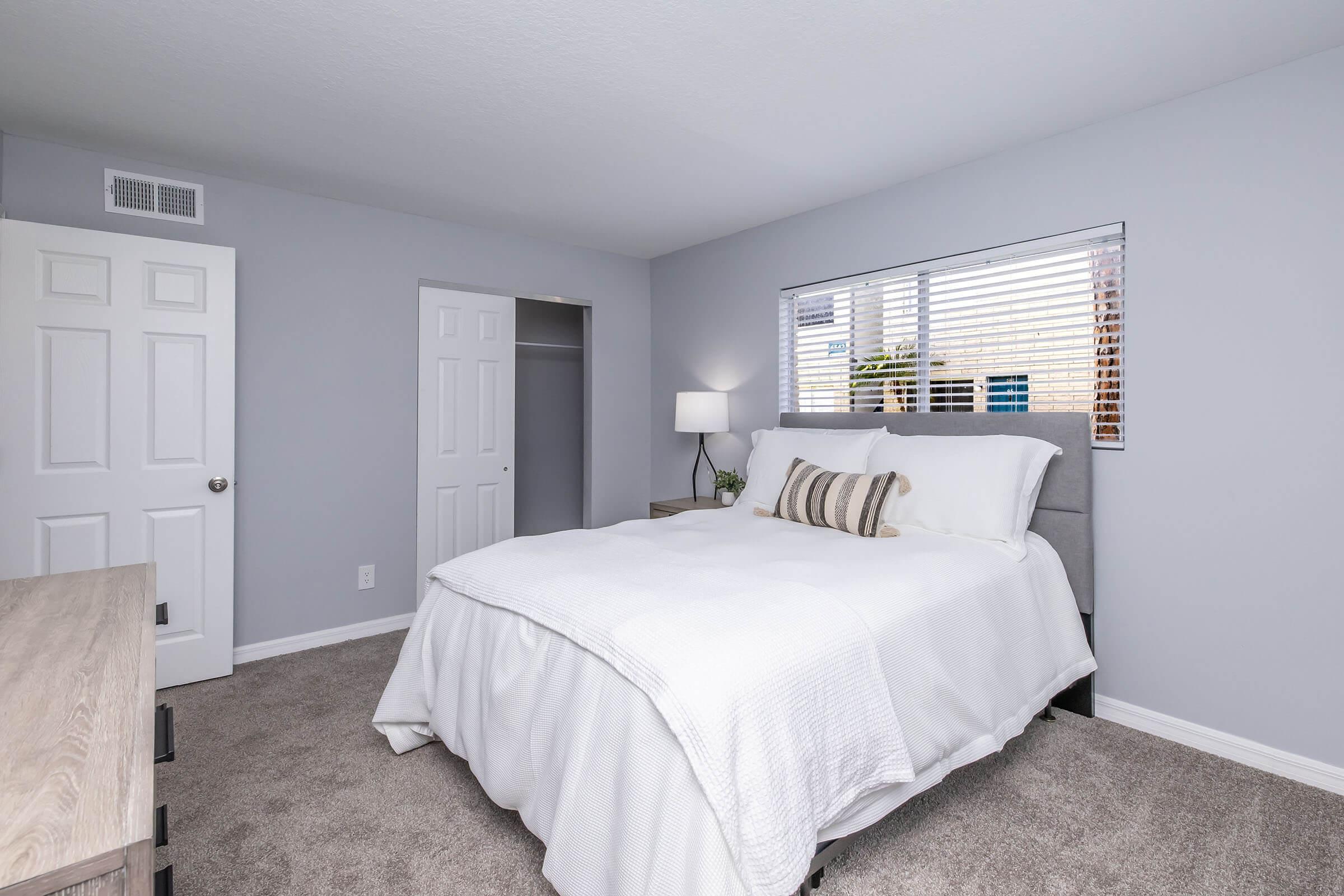 A cozy bedroom featuring a light gray color scheme, a neatly made bed with white bedding and decorative pillows, a bedside lamp on a wooden nightstand, a window with blinds allowing natural light, and a closet with double doors. The carpet is plush, enhancing the room's comfort.