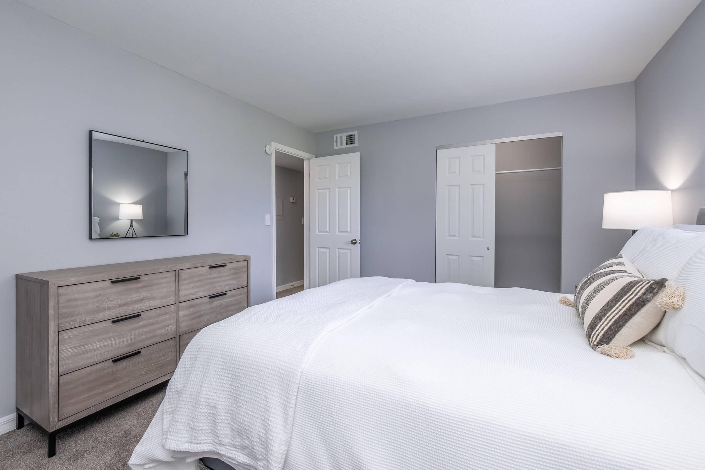 A modern bedroom featuring a neatly made bed with white bedding and a decorative pillow. There is a wooden dresser with drawers, a mirror on the wall, and a doorway leading to a closet. The walls are painted in a light gray color, and the room is well-lit, creating a clean and inviting atmosphere.