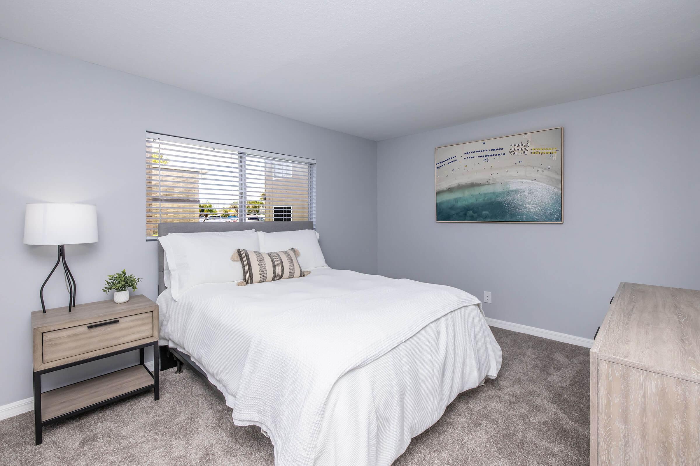 A bright and modern bedroom featuring a queen-sized bed with white bedding, a small nightstand with a lamp and a potted plant, and a wooden dresser. Natural light streams in through a window with blinds, while a piece of artwork depicting a serene beach scene hangs on the wall.