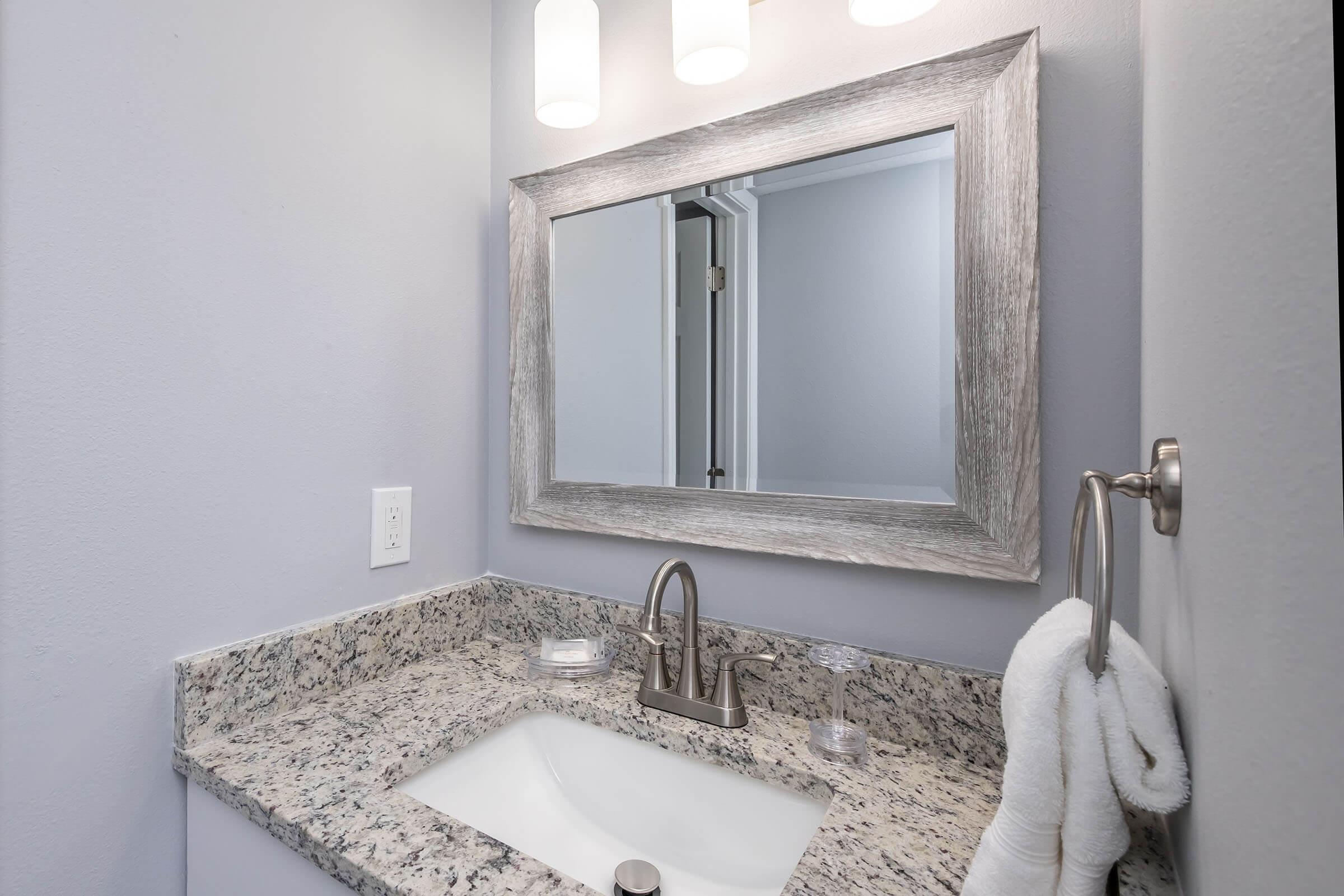 A modern bathroom featuring a granite countertop with a sink. Above the sink, a stylish mirror framed in light gray wood is illuminated by three pendant lights. A towel hangs on a chrome holder beside the sink, and the wall is painted in a soft gray color. The overall design is sleek and contemporary.