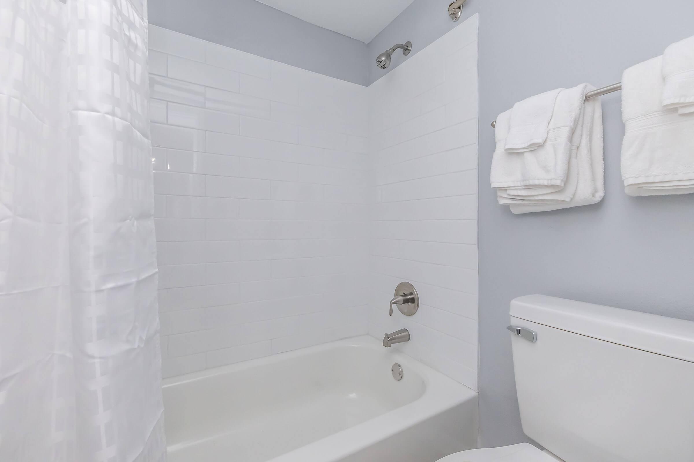 A clean bathroom featuring a white bathtub with a shower curtain, white tile walls, and a toilet. Fresh, folded white towels are neatly displayed on a towel rack beside the shower. The wall color is light gray, creating a calm and spacious appearance.