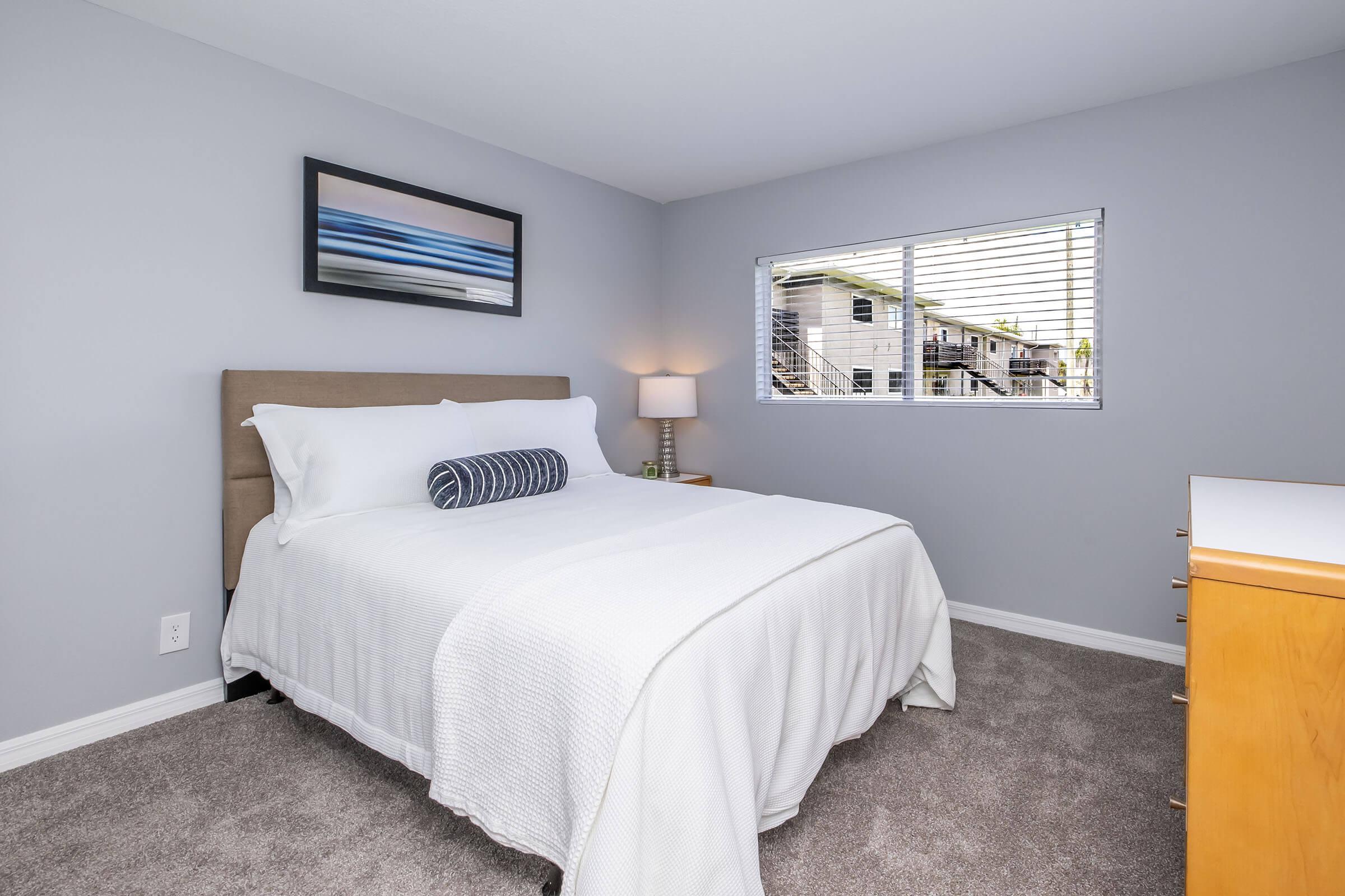 A cozy bedroom featuring a queen-sized bed with white linens and a decorative pillow. A bedside lamp illuminates the space, and a window with blinds lets in natural light. The walls are painted in a soft gray, and there's a wooden dresser against one wall, creating a calm and inviting atmosphere.