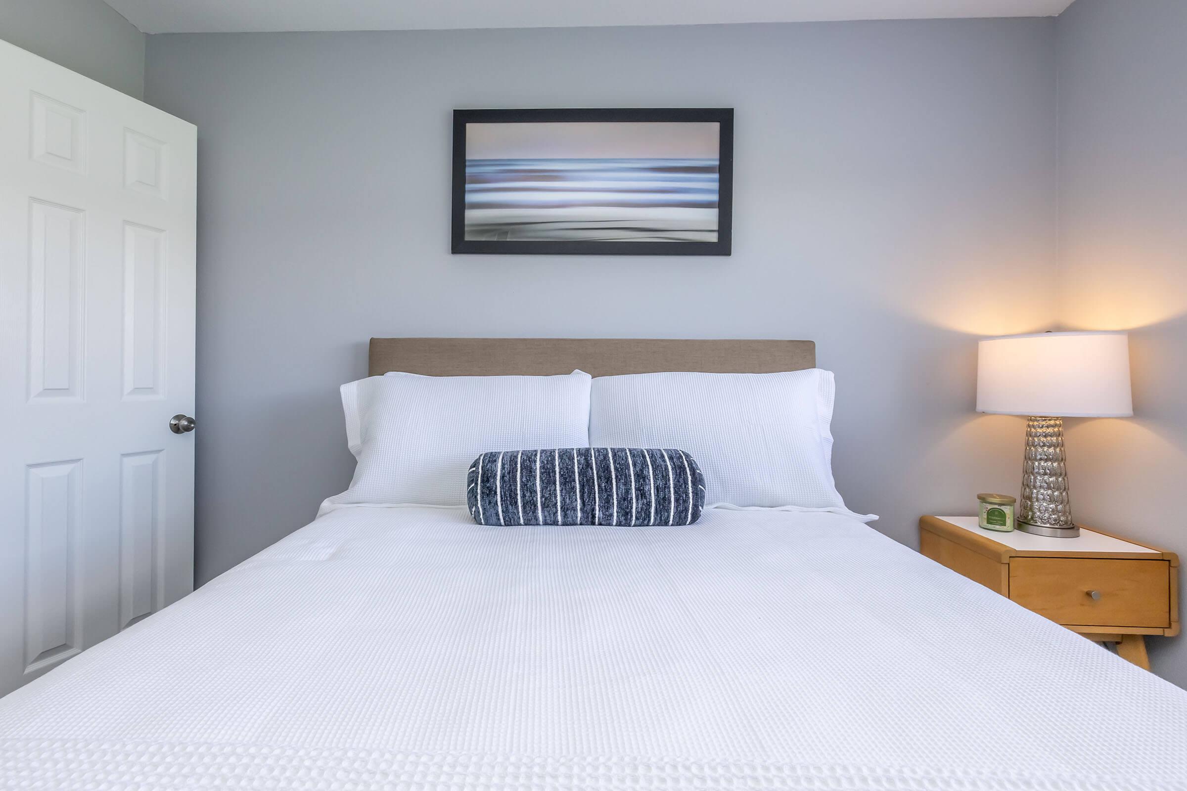 A neatly made bed with white linens and a decorative blue-striped bolster pillow. A nightstand with a small candle and a lamp stands beside the bed, and a landscape artwork hangs on the wall above. The room features light gray walls and a closed white door in the background.