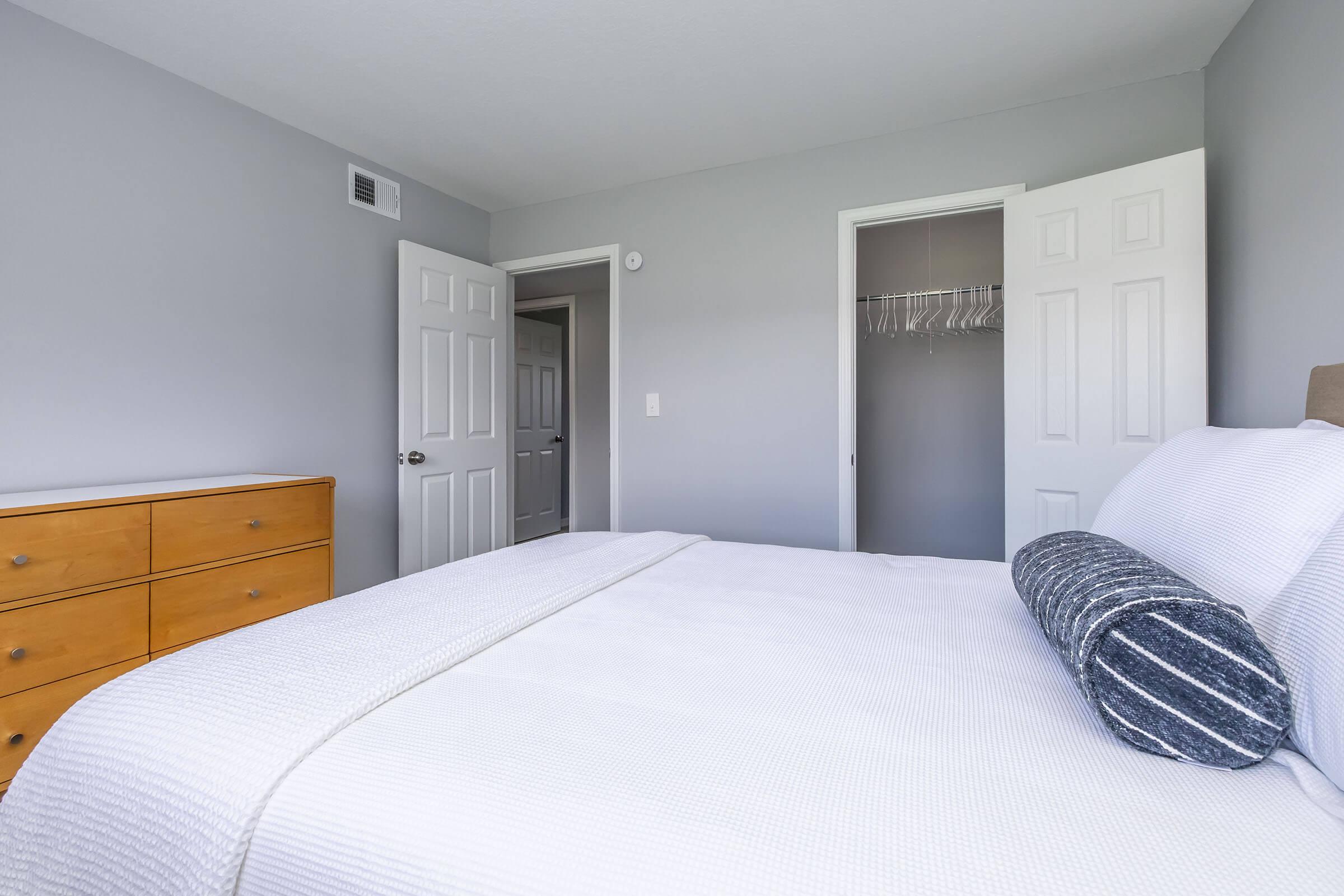 A modern bedroom featuring a neatly made bed with a white comforter and a decorative pillow. To the side, there is a wooden dresser with multiple drawers. The walls are painted in a light gray color, and there are two open doors leading to closets, revealing additional space. Natural light fills the room, creating a bright and inviting atmosphere.